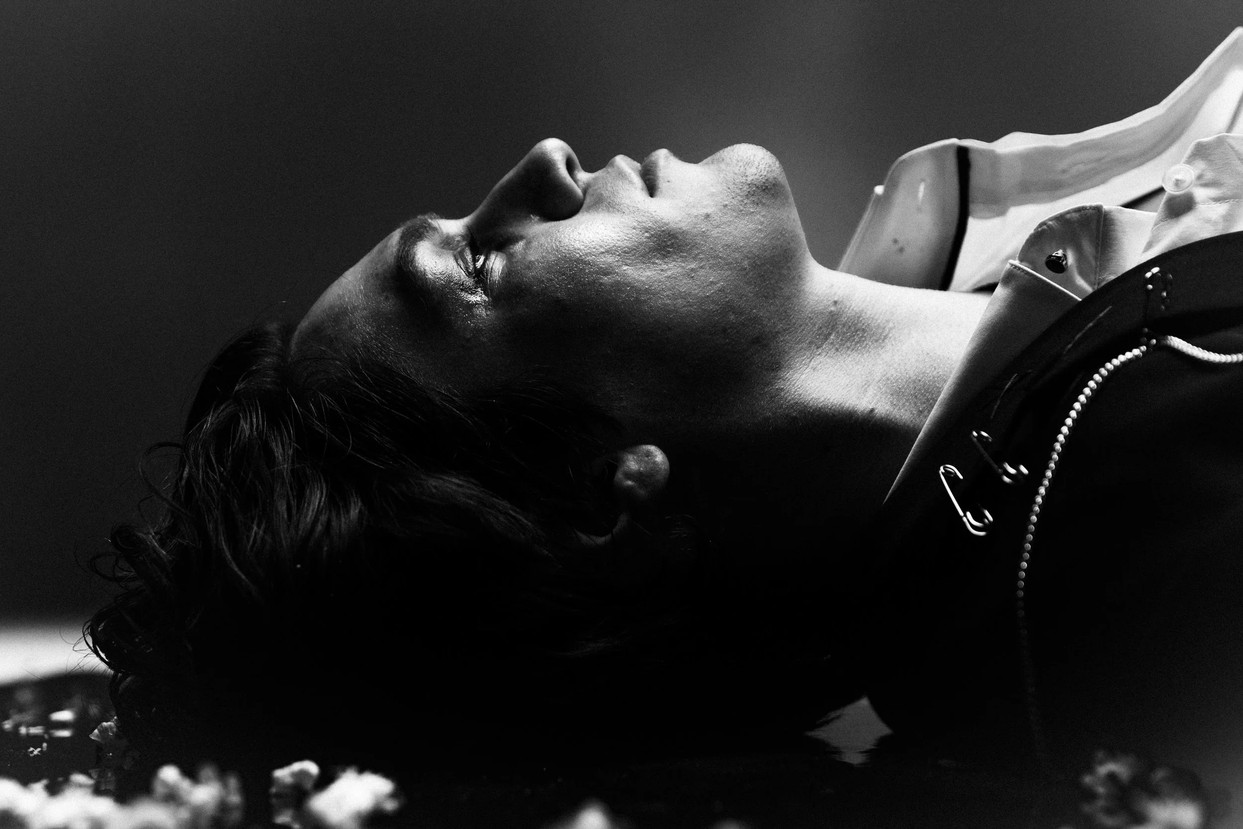 Black and white photo of a woman lying on her back with her eyes closed, wearing a jacket and a necklace, with scattered popcorn on a surface below.