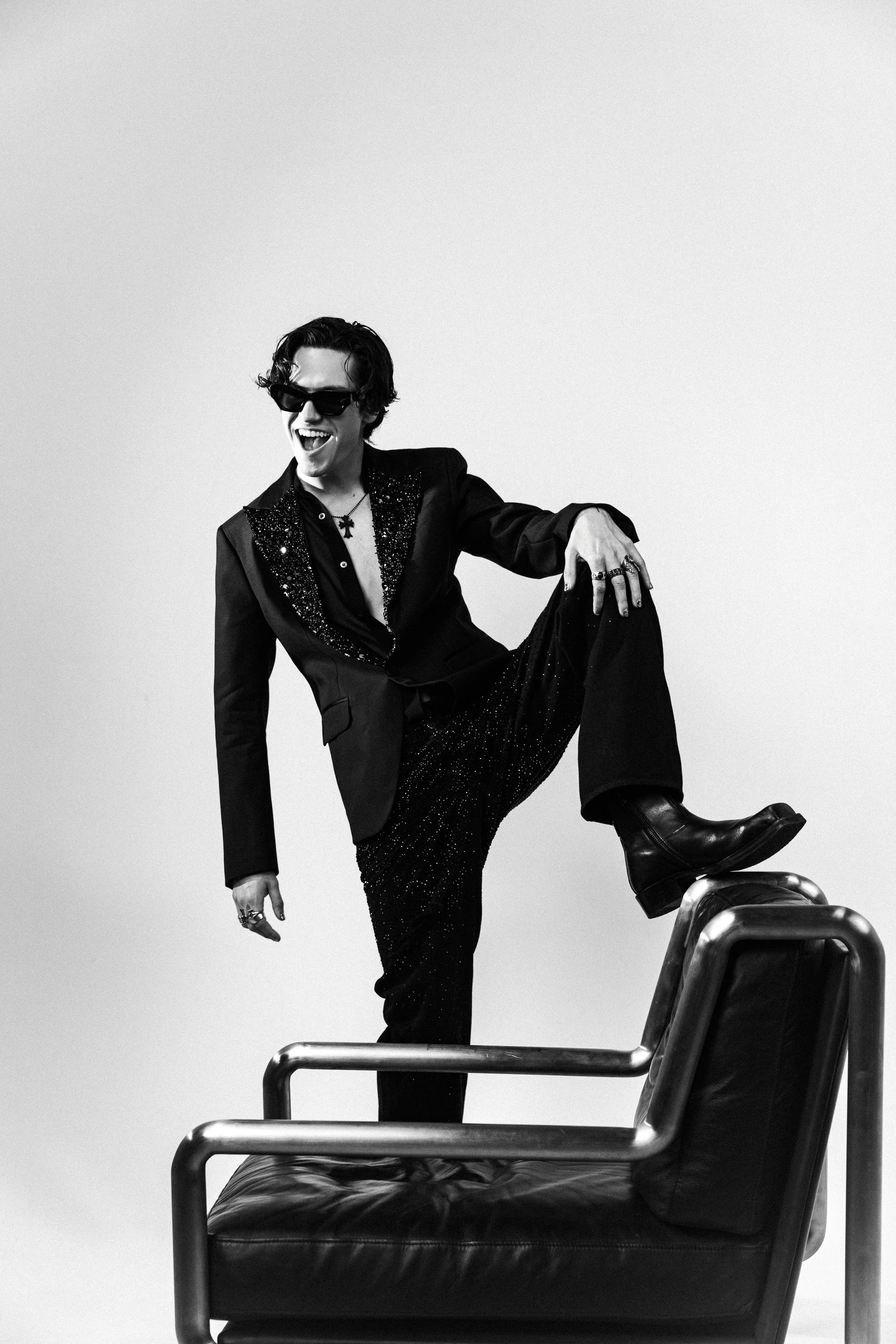 Black and white photo of a man with dark sunglasses, wearing a glittery black suit, laughing or smiling, with his right leg propped up on a barstool, against a plain light background.
