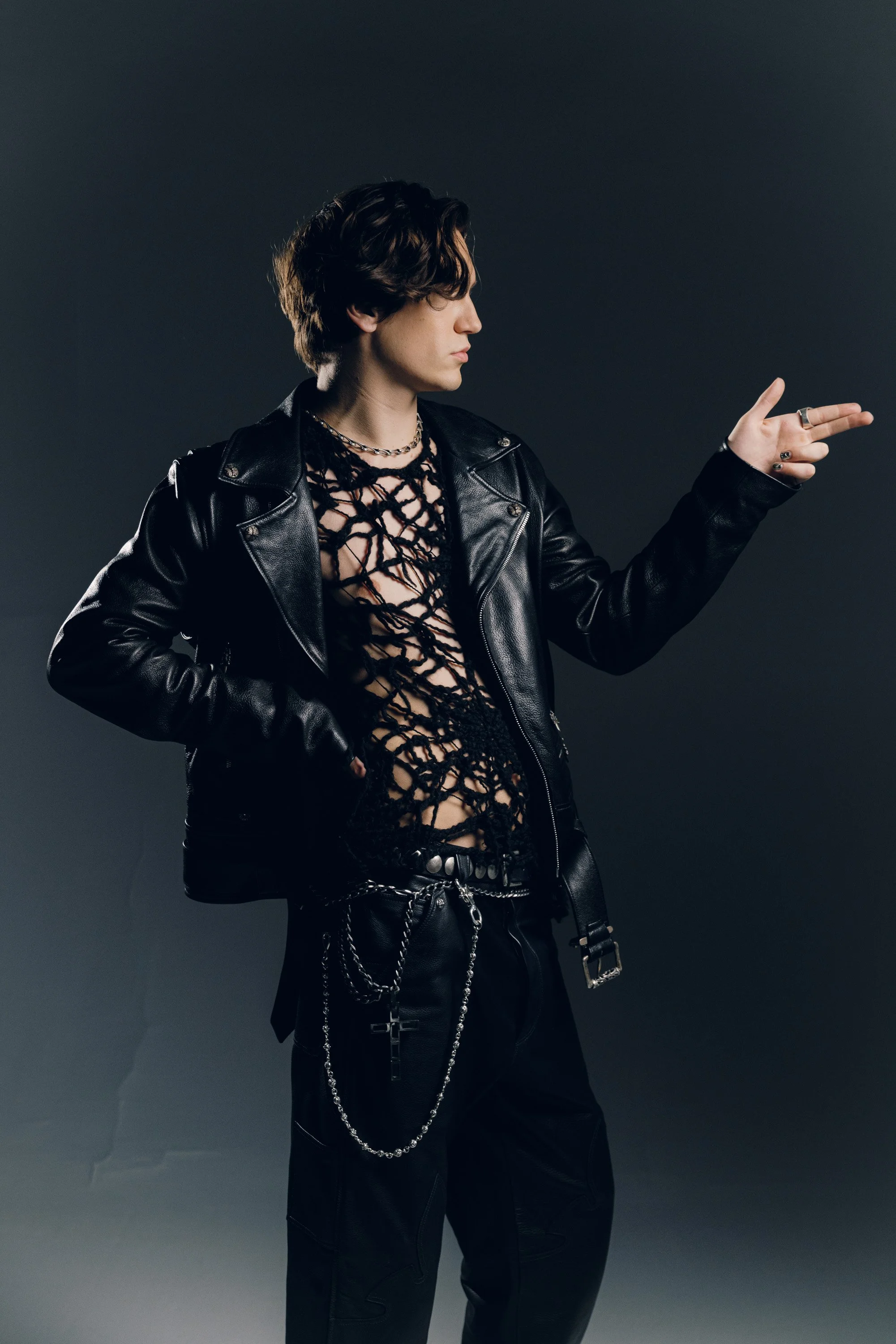 Young man with wavy dark hair wearing a black leather jacket, fishnet shirt, and jewelry, posing against a dark background, making a peace sign with his right hand.