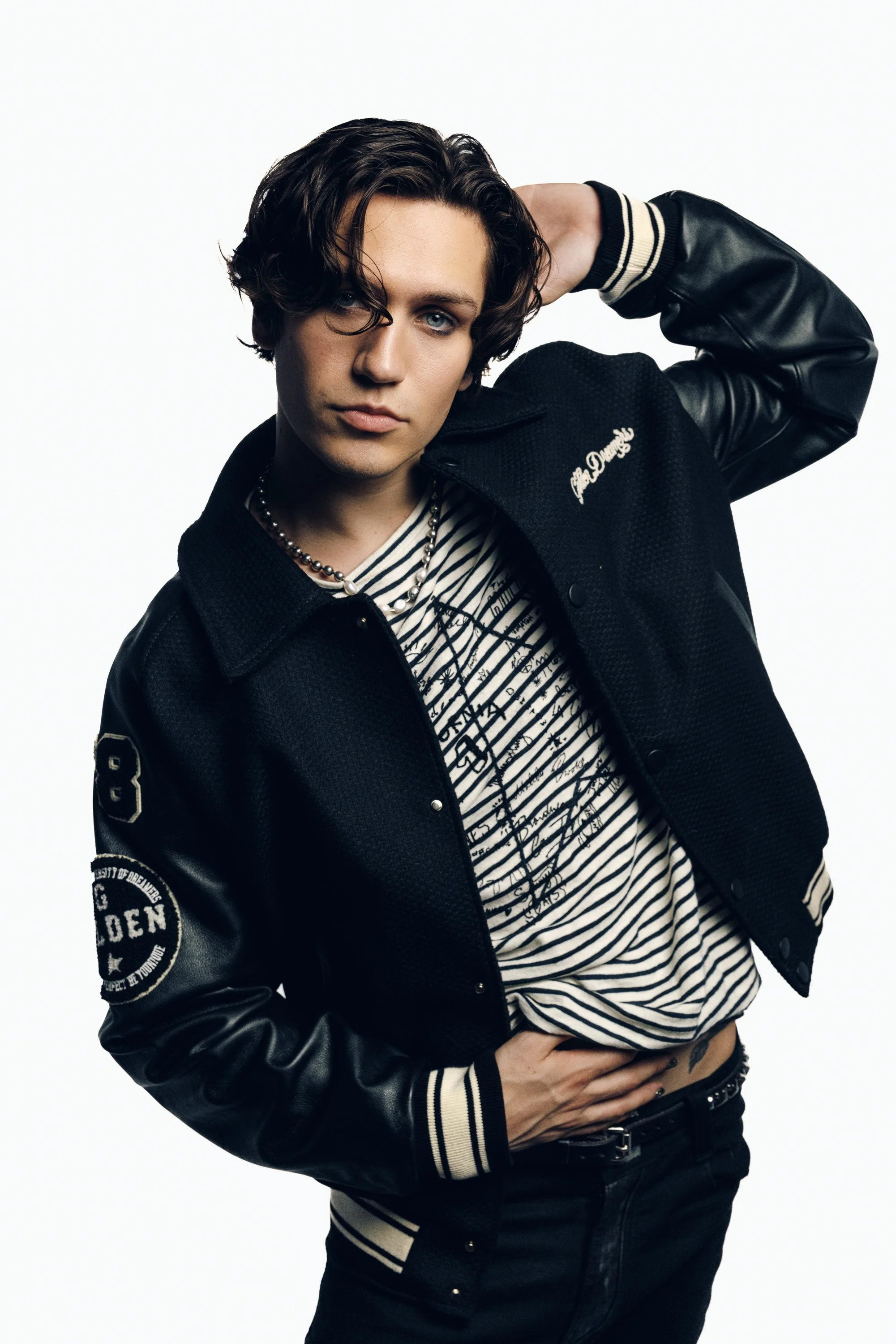 Young man with dark wavy hair and striking blue eyes posing with one hand behind his head, wearing a black and white striped shirt, a black varsity jacket, and black pants against a white background.