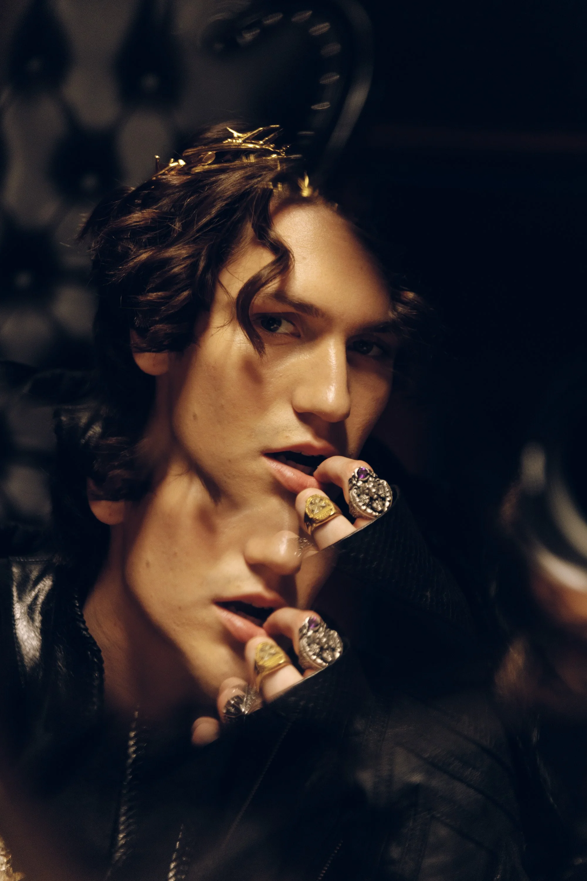 Close-up of a young man with long, wavy dark hair, wearing multiple rings and a black leather jacket, posing against a dark background.