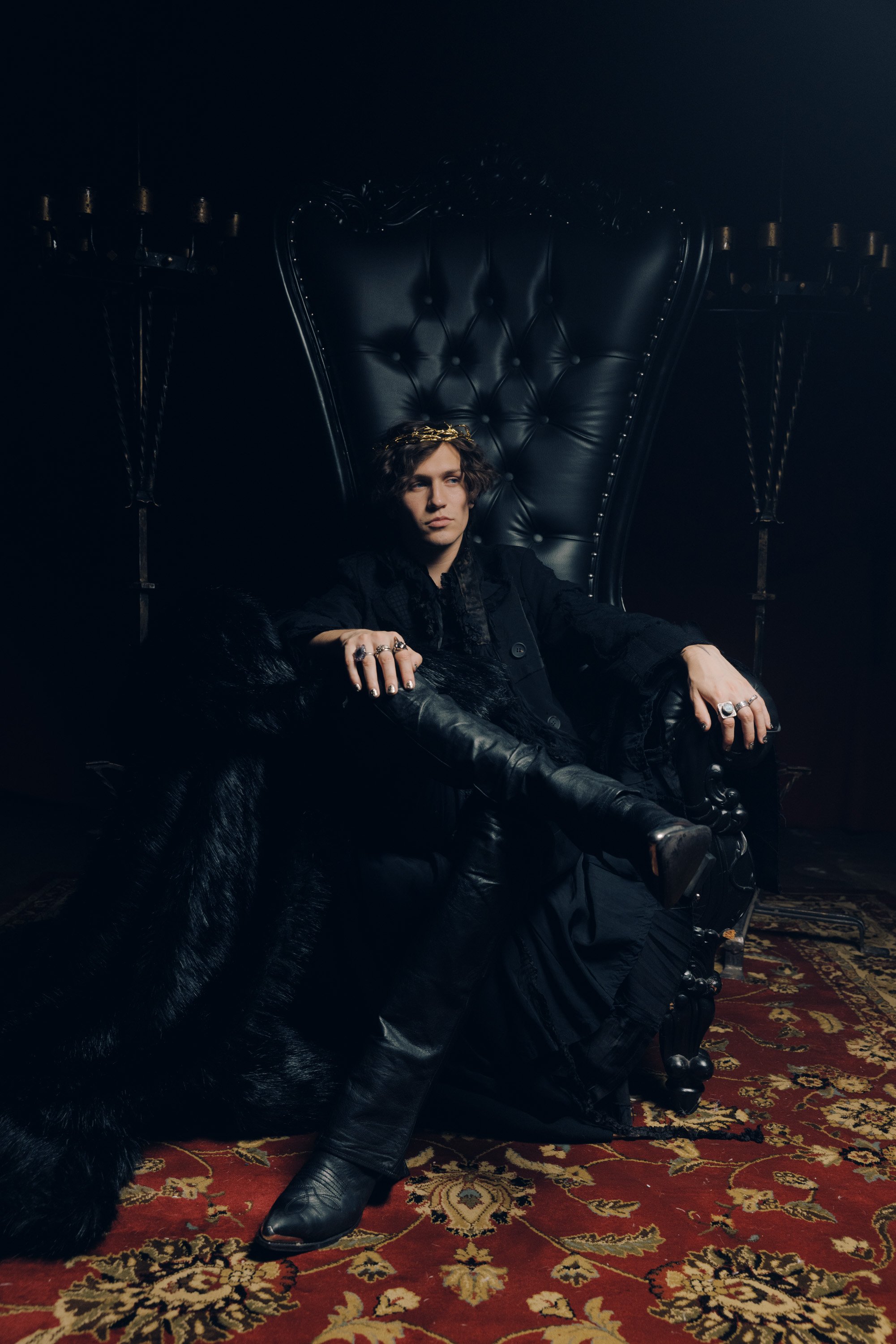 Person sitting on an ornate black leather throne with a high backrest, in a dark room with candelabras on either side, wearing black clothing and jewelry, and a gold headpiece, sitting on a patterned carpet.