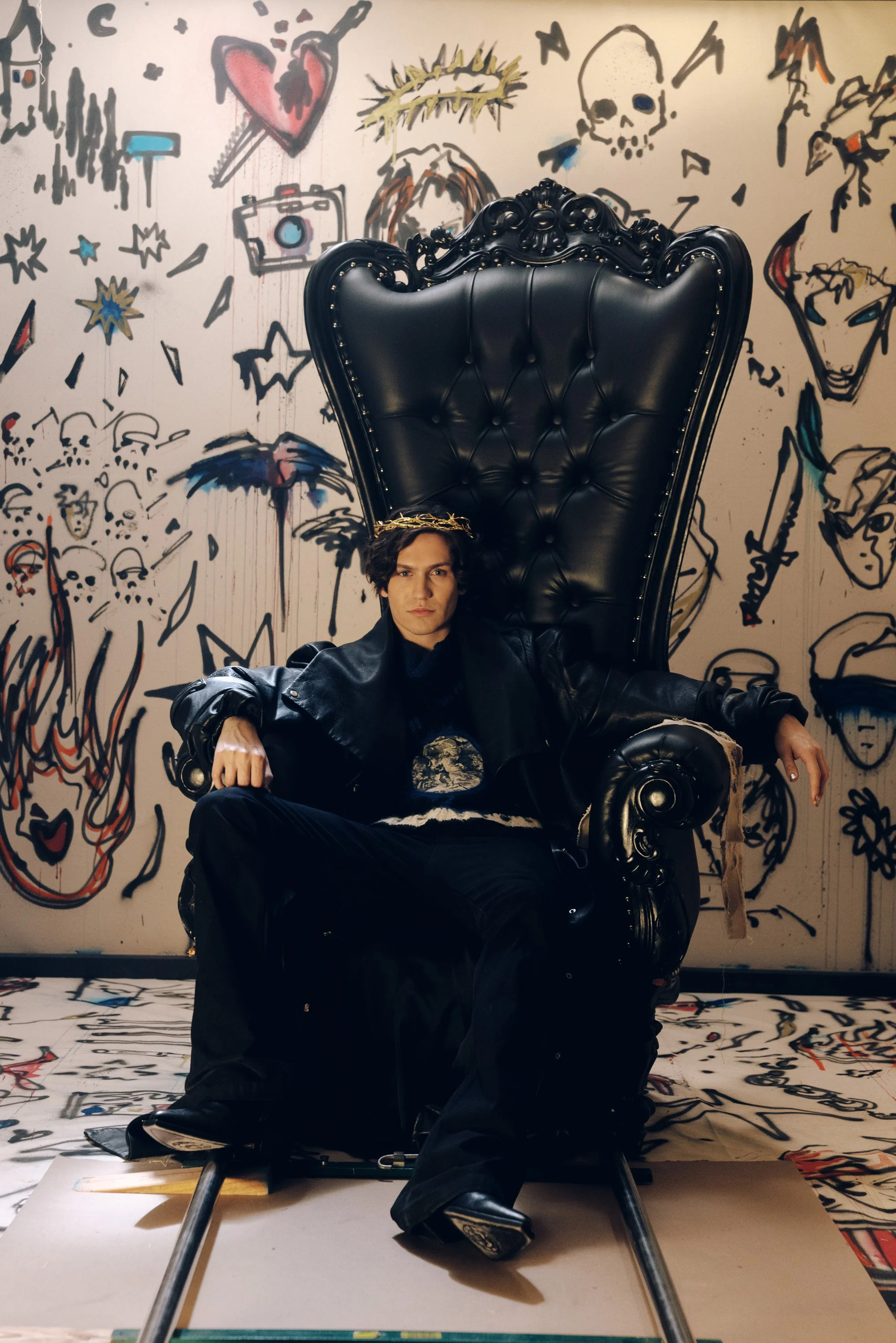 A person sitting on a large, ornate black throne with gold details, in front of a wall covered with graffiti-style art, including a skull, a heart with a dagger, and various abstract figures and faces.