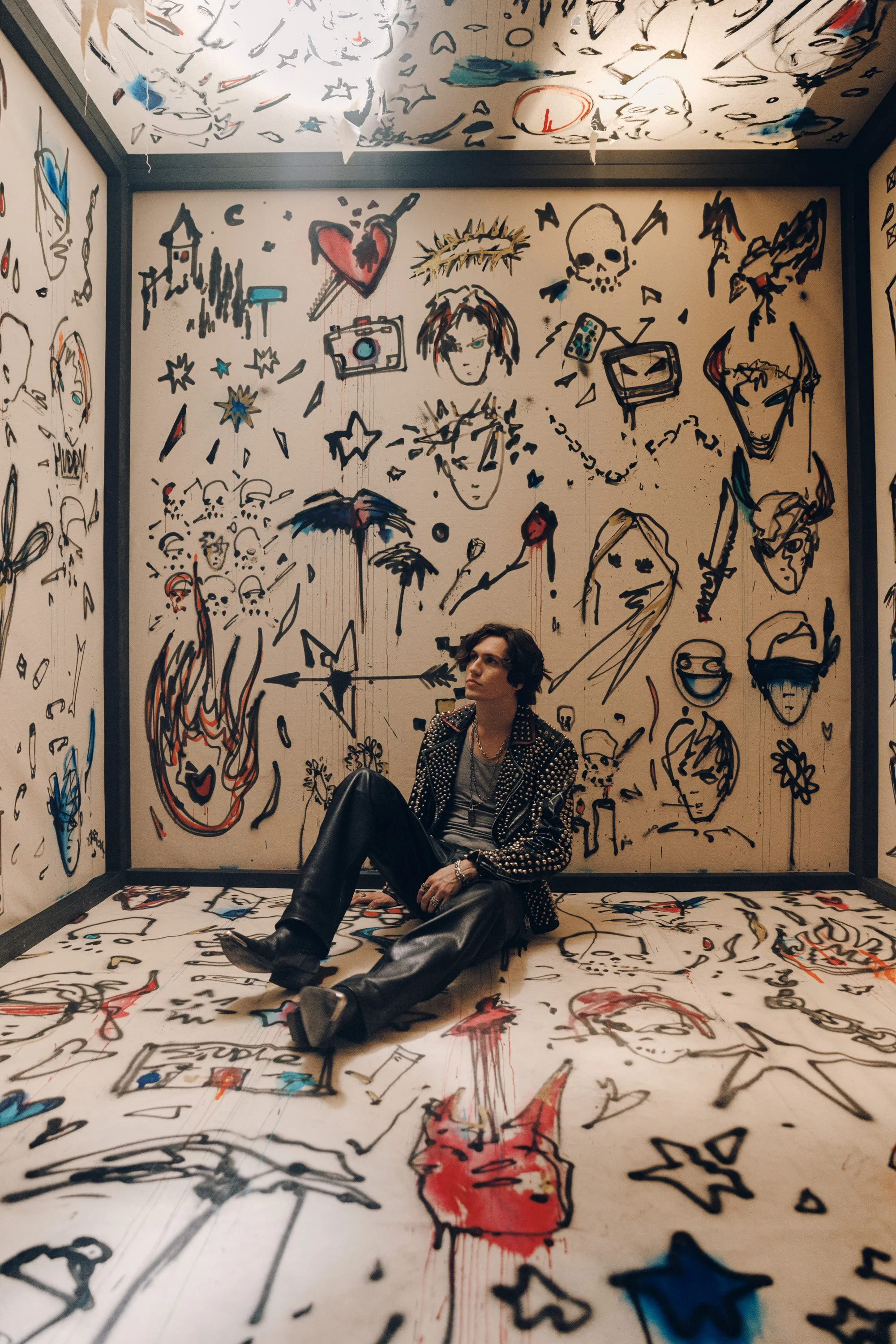 A young woman with dark hair, dressed in a studded black jacket, gray shirt, and leather pants, sitting on a floor covered with graffiti-style drawings and illustrations. She is relaxing against a wall that is also adorned with similar graffiti artwo