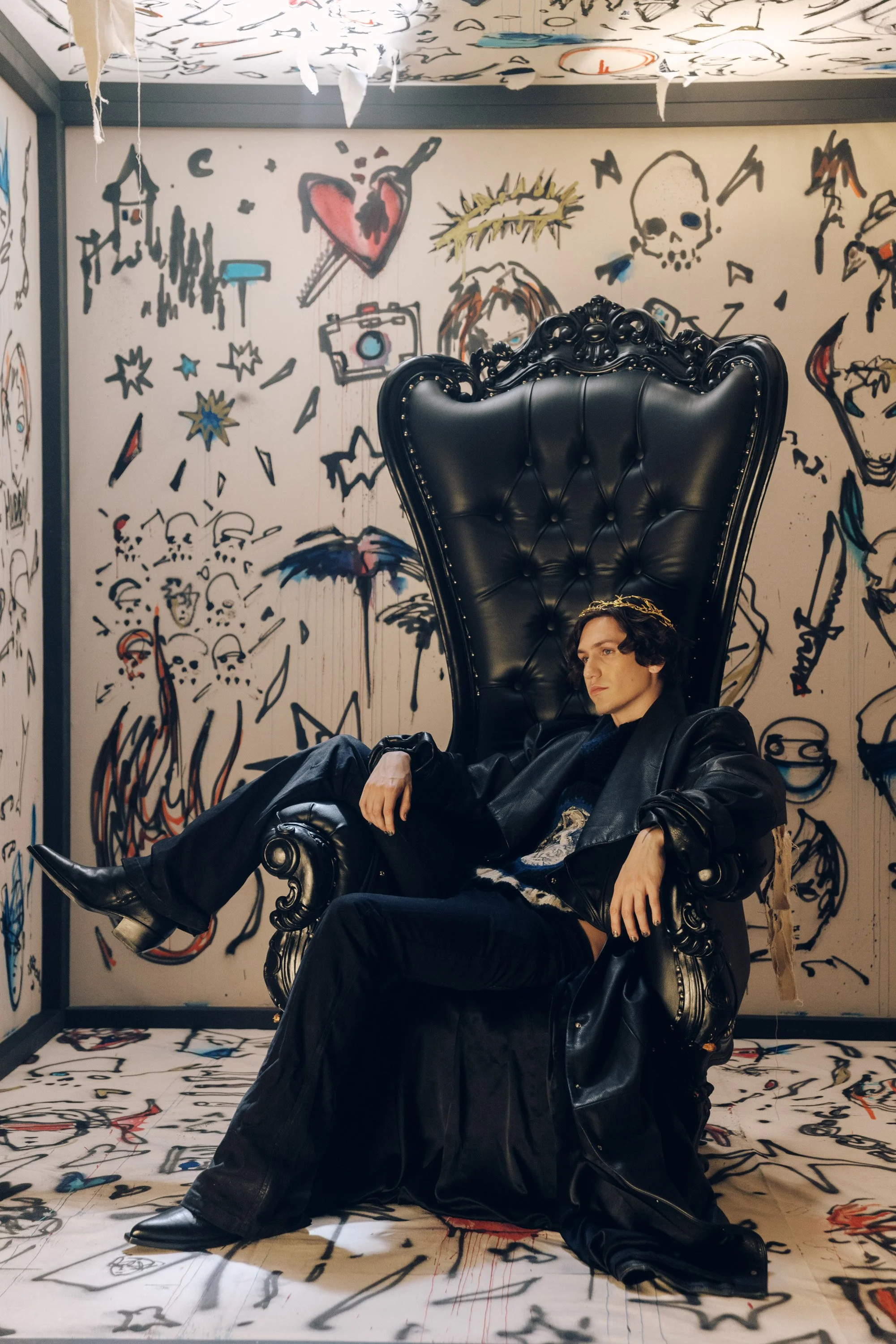 A person with short dark hair, dressed in black, sitting in a large, ornate black leather armchair with a high back. The background features a white wall with colorful graffiti-style drawings including a heart with a dagger, skulls, stars, faces, and