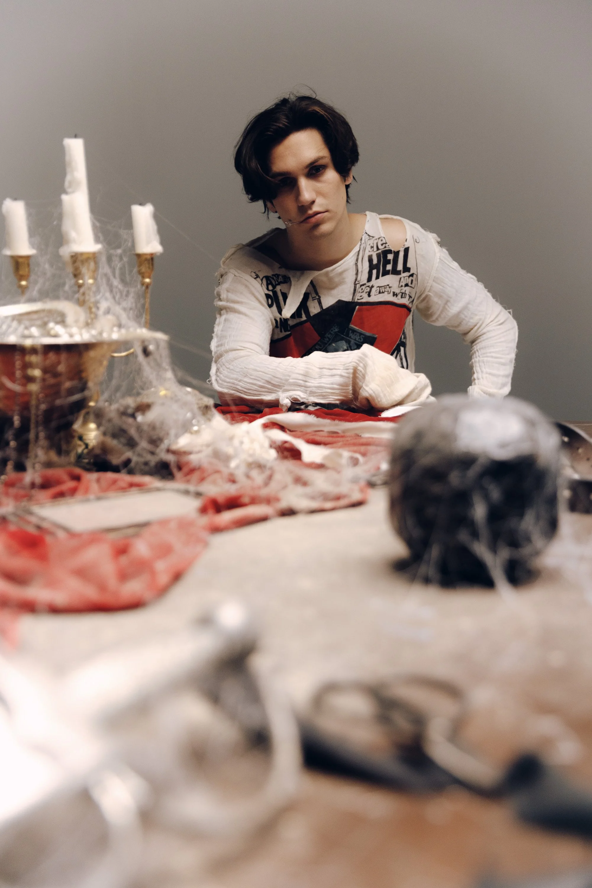 A person with dark hair, wearing a distressed white sweatshirt with bold black and red text, sitting at a table with Halloween-themed decorations including candles, cobwebs, and faux spiderwebs, with a neutral background.