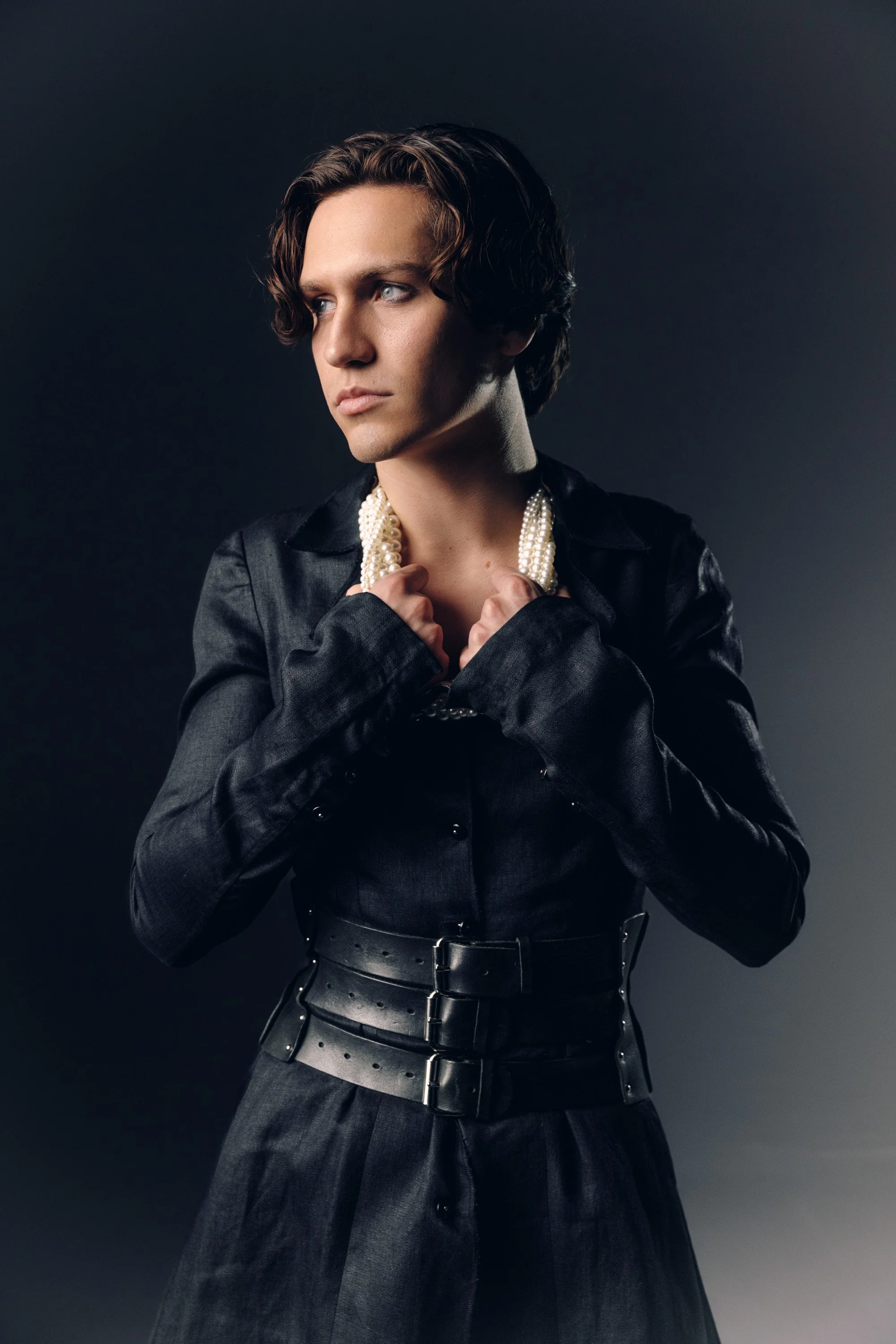 A person with short, wavy dark hair, wearing a black leather outfit with a belt, holding a multi-strand pearl necklace and looking away from the camera against a dark gray background.