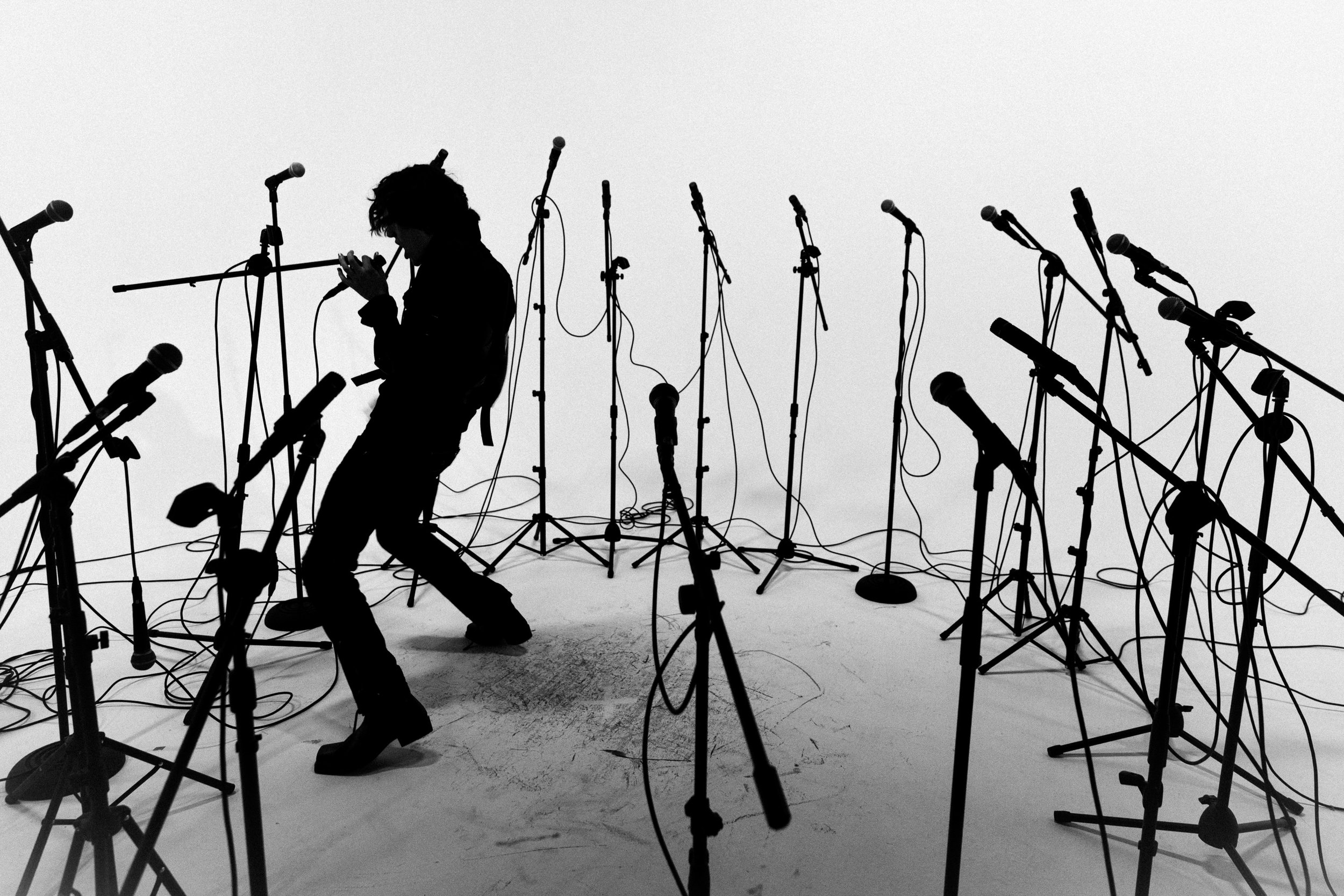Silhouette of a person holding a microphone amidst numerous stand microphones in a minimalistic, black-and-white setting.
