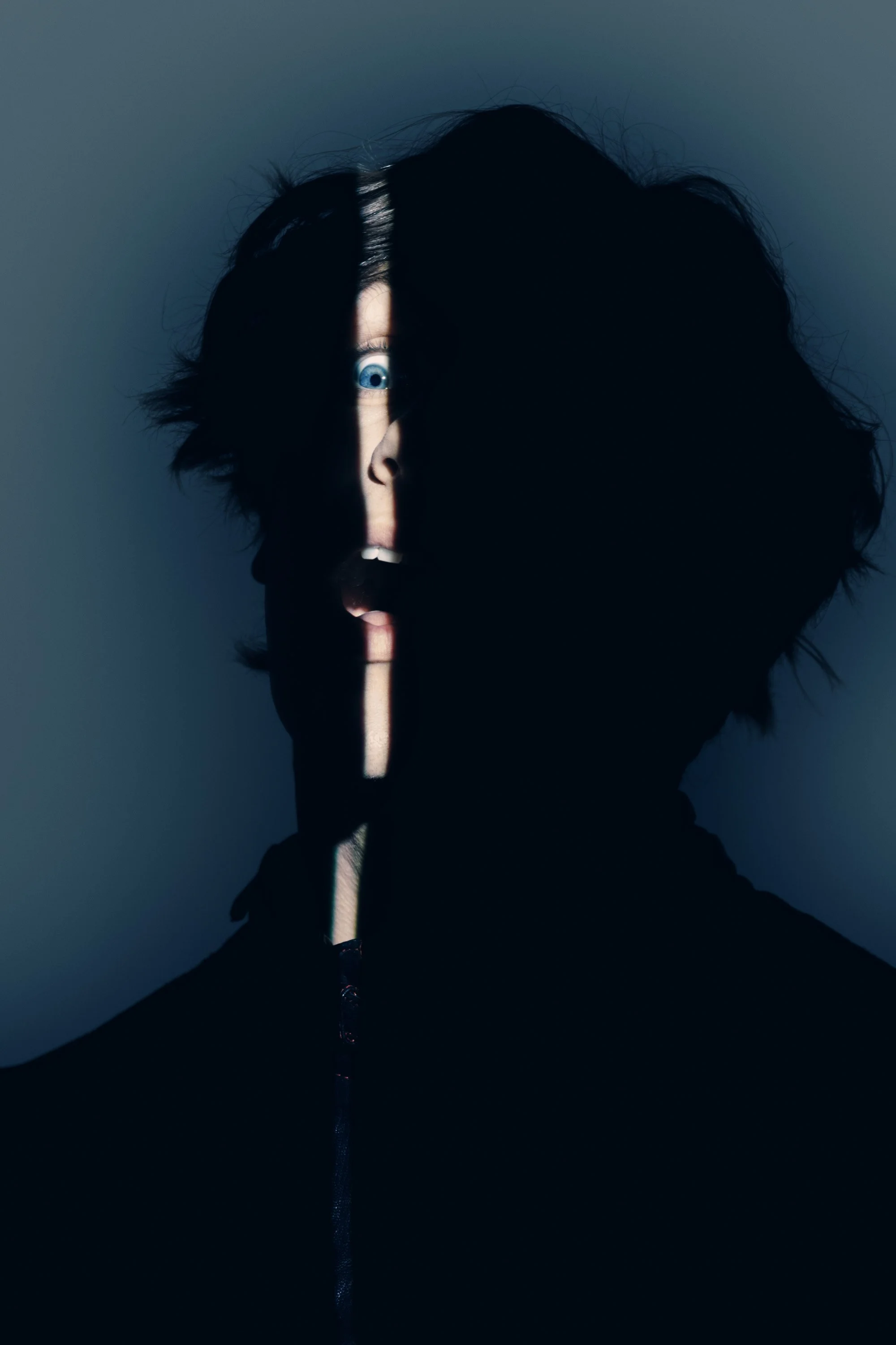 A person facing a wall with a shadow cast across half of their face, revealing one eye and an open mouth, with dark hair and a dark background.