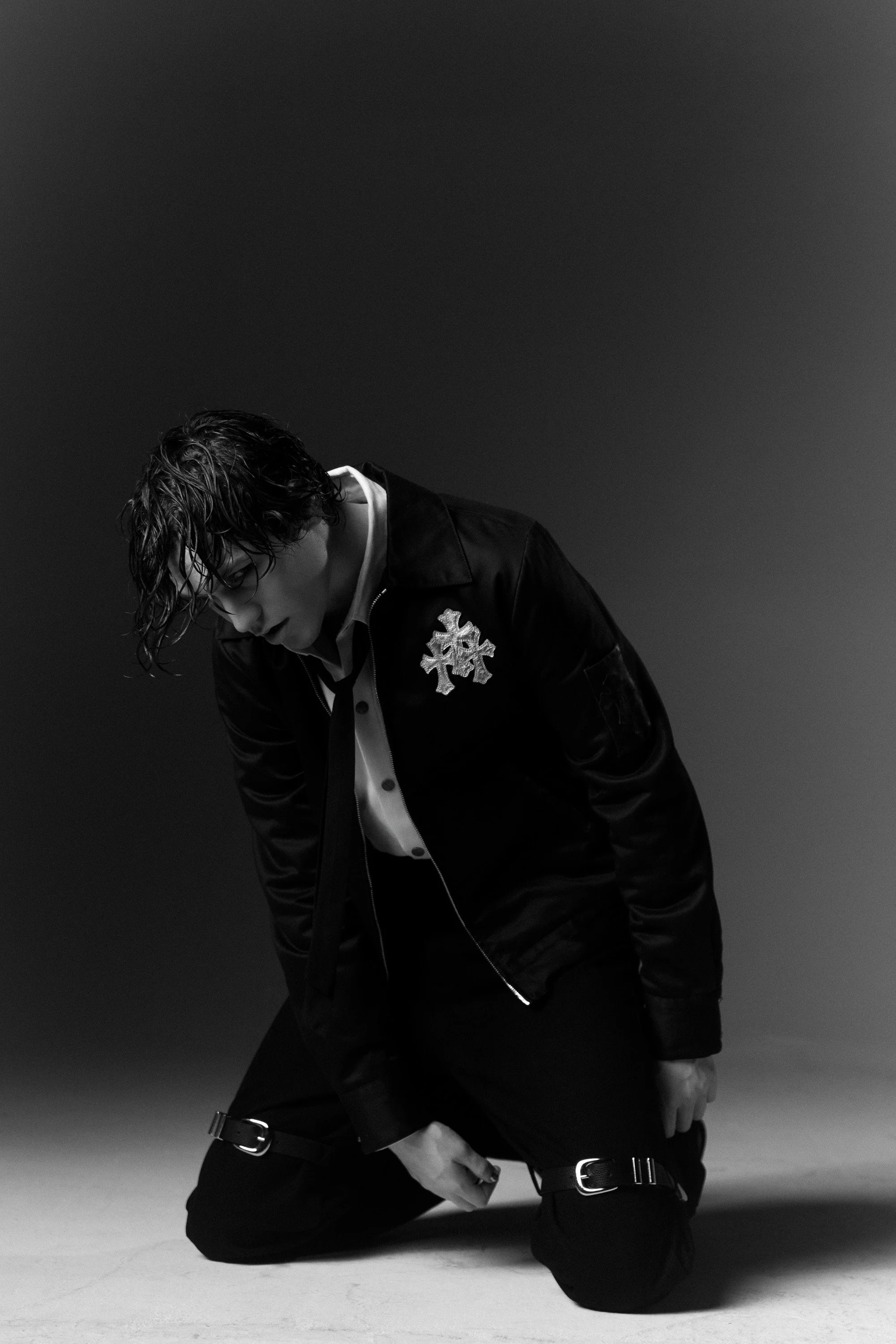 Black and white photo of a young man with wet hair, wearing a jacket with a snowflake patch, kneeling on the ground with a serious expression.