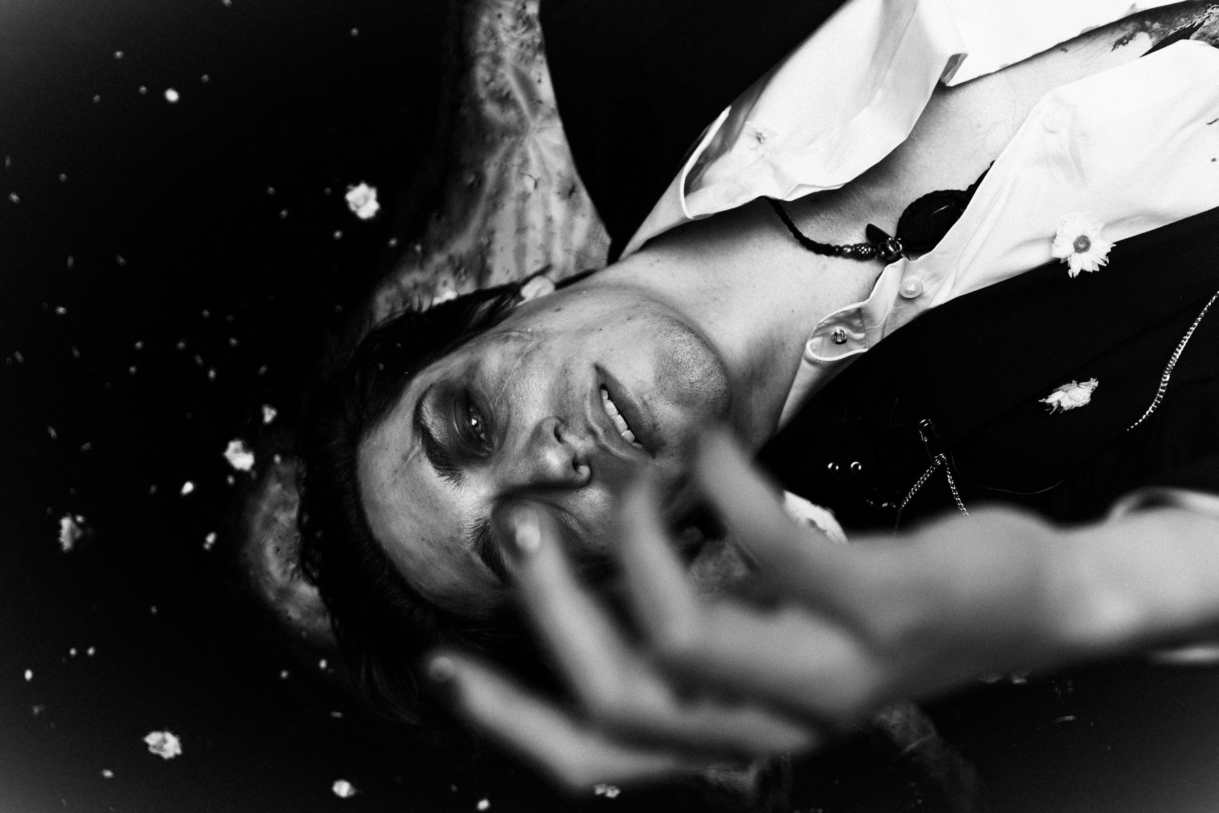 A person lying down, close-up of their face, wearing a white shirt with a necklace, and surrounded by small flowers or petals.