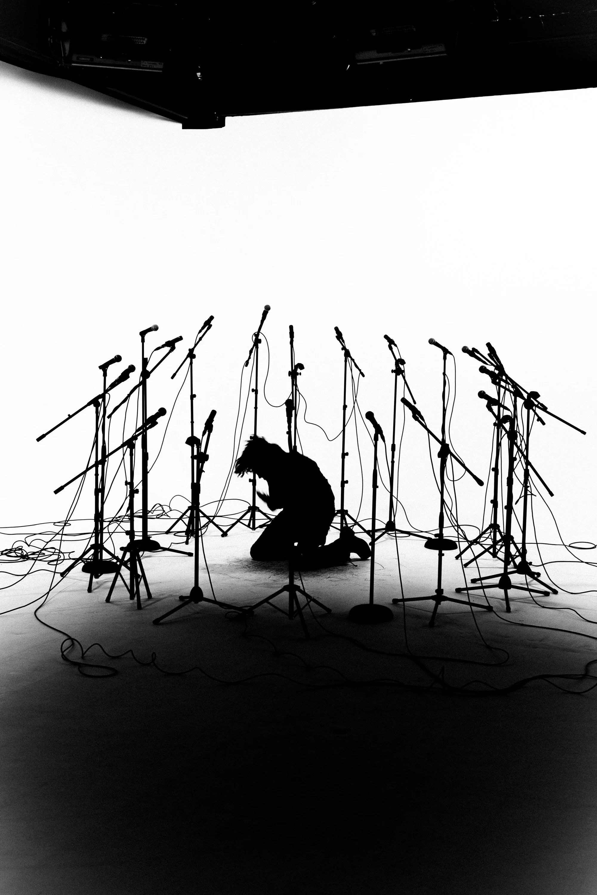 A black and white photo of a stage with many microphone stands and cords, with a silhouette of a person kneeling among the equipment, viewed from a low angle against a bright background.