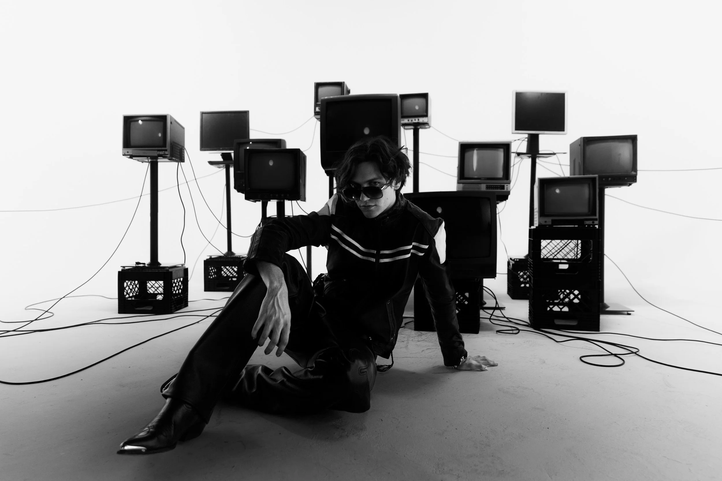 A person with sunglasses and tousled hair crouches on the floor surrounded by vintage monitors and wires in a monochrome studio setting.