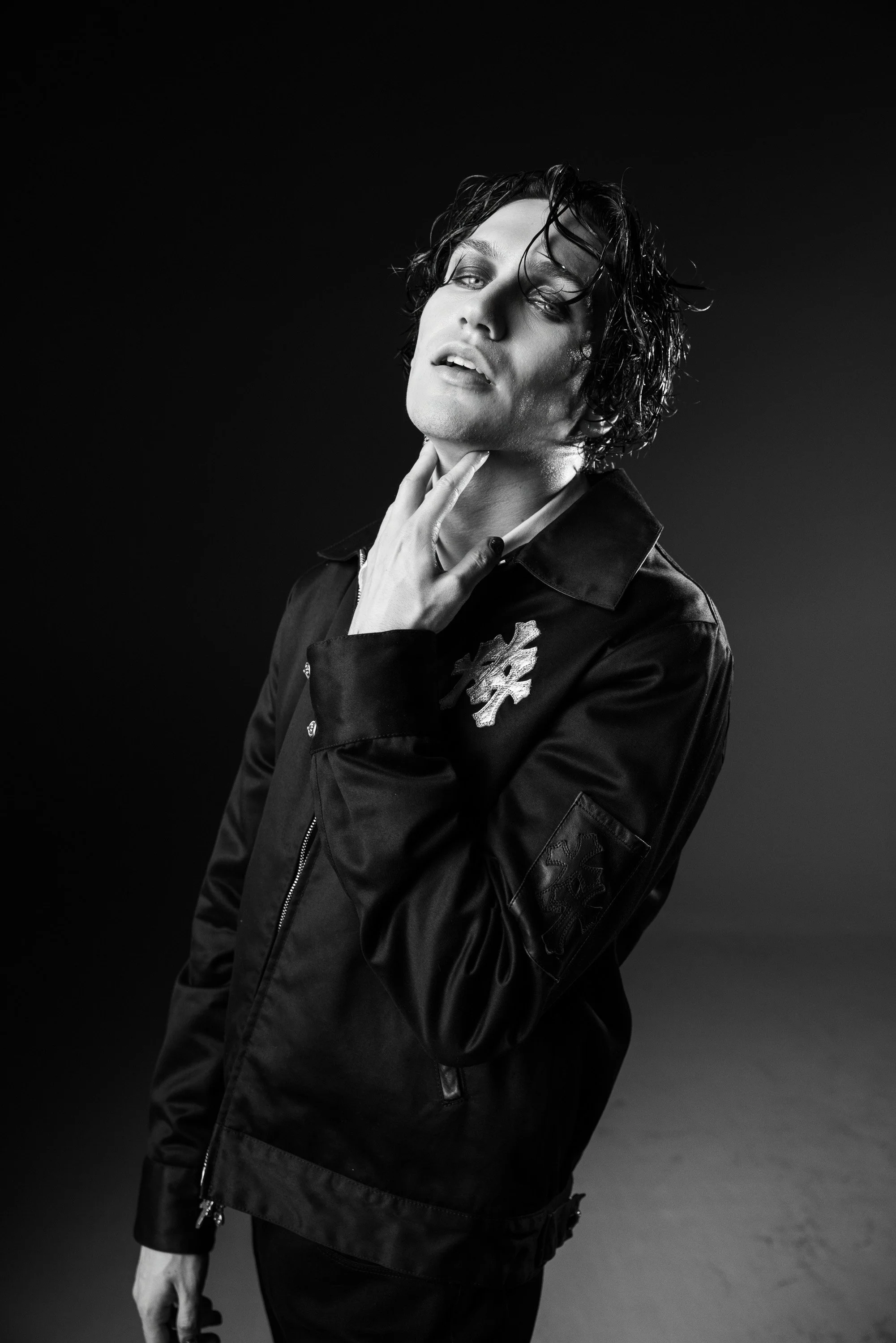 Black-and-white portrait of a person with wet, curly hair, wearing a dark jacket with floral embroidery, touching their neck with one hand, against a black background.