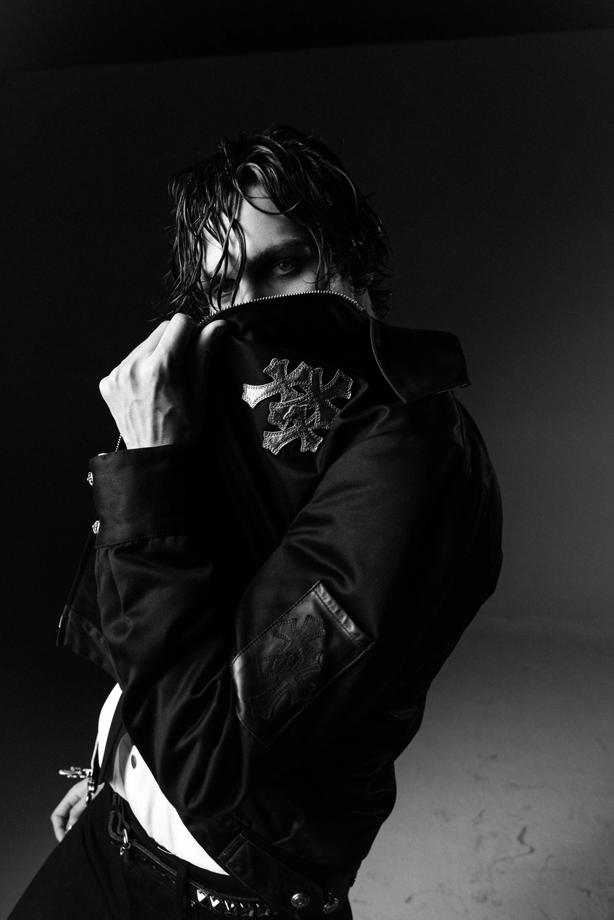 A person with wet, tousled hair partially covering their face, wearing a dark jacket with decorative patches, holding the collar up over their mouth and nose, in a black and white photograph against a dark background.