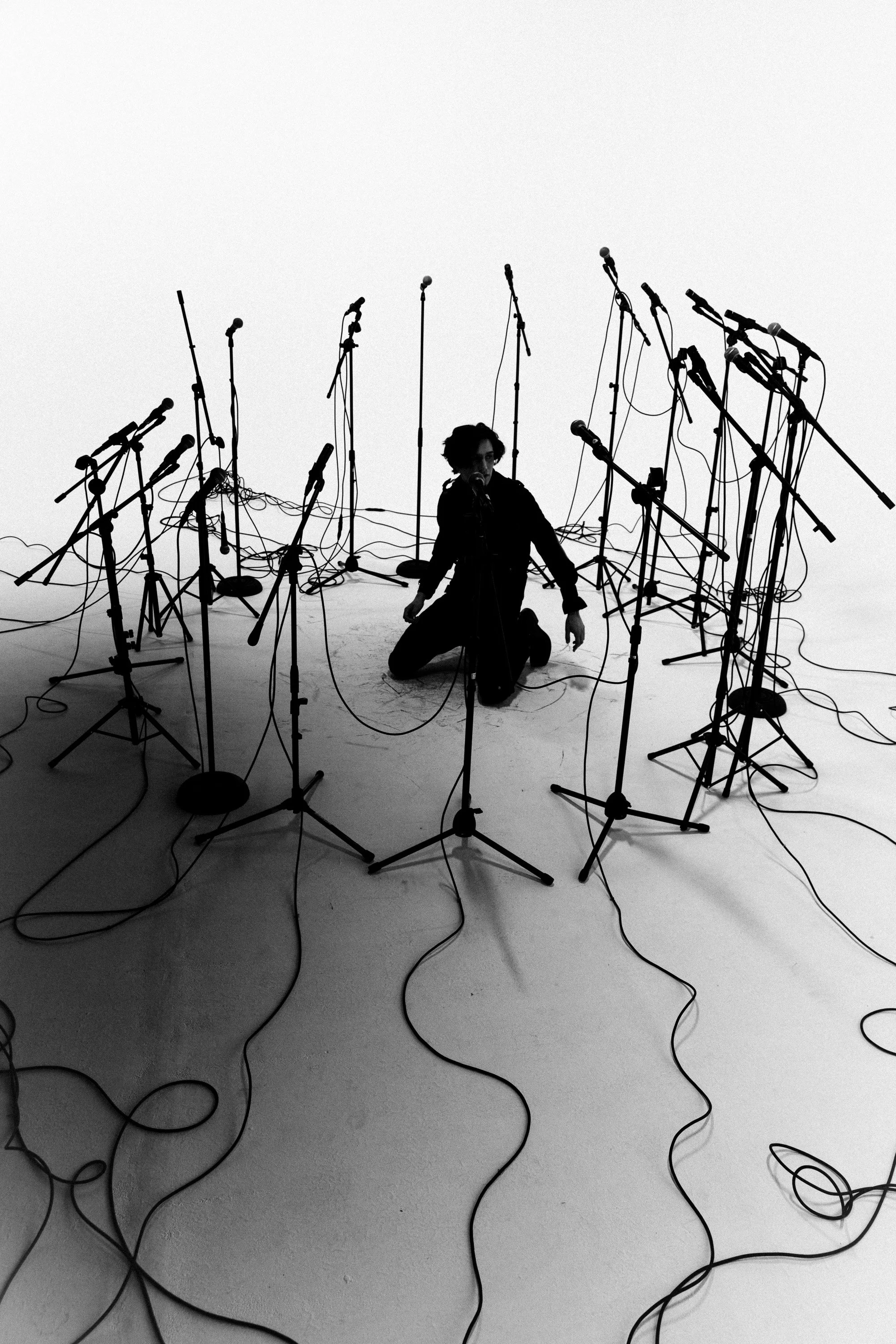 A person kneeling on the floor surrounded by many microphone stands with wires, against a plain white background.