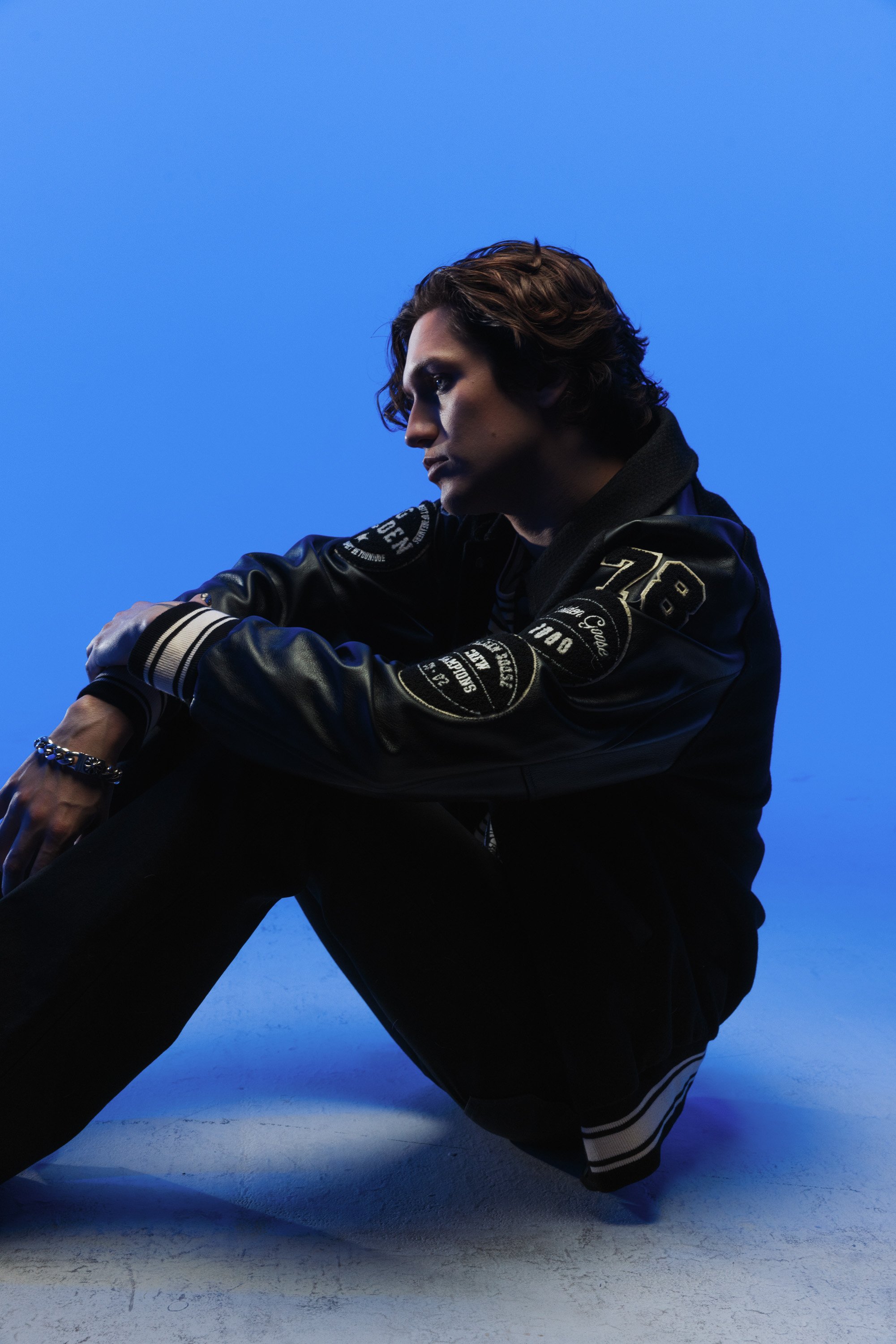A person with wavy brown hair sitting on the floor against a blue background. They are wearing a black jacket with patches and lettering, and black pants with white stripes at the bottom, with their arms resting on their knee.