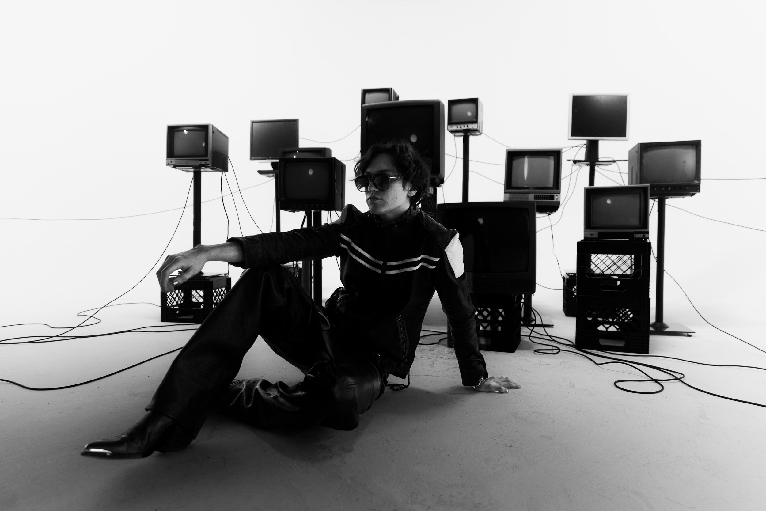 A person wearing sunglasses and a black leather jacket sitting on the floor in front of a white background with multiple vintage televisions on stands behind them, connected by wires.