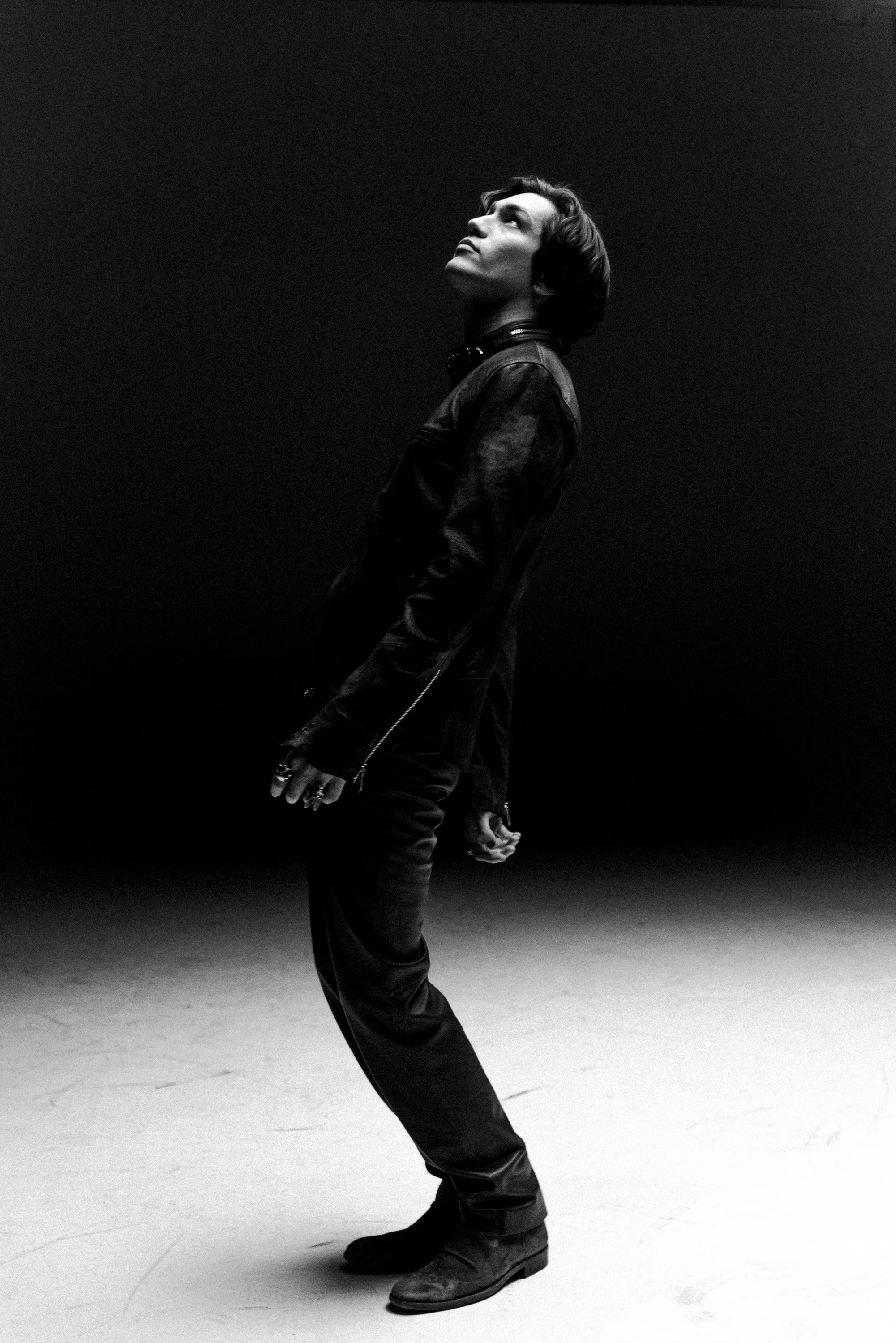 Black and white photo of a woman standing in a studio, looking upward with a thoughtful expression. She is wearing a leather jacket, black pants, and boots, with headphones around her neck. The background is plain and dark.