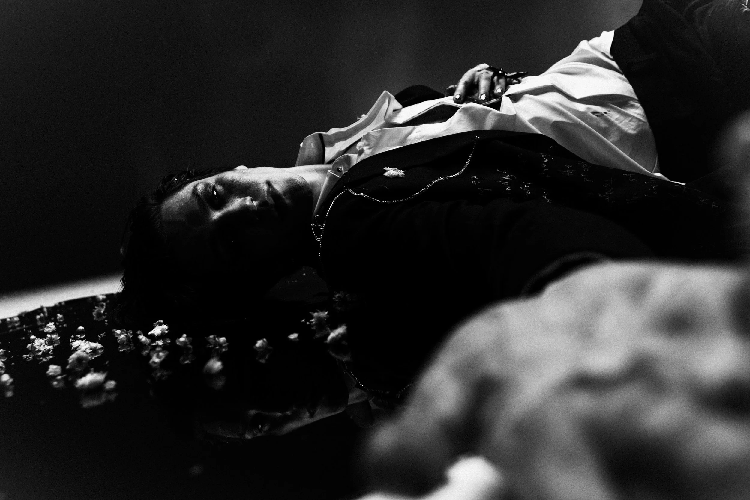 Black and white photo of two individuals lying on a surface covered with scattered white flower petals, with one person visible only from the shoulders up, lying on her side facing the camera with a calm expression.