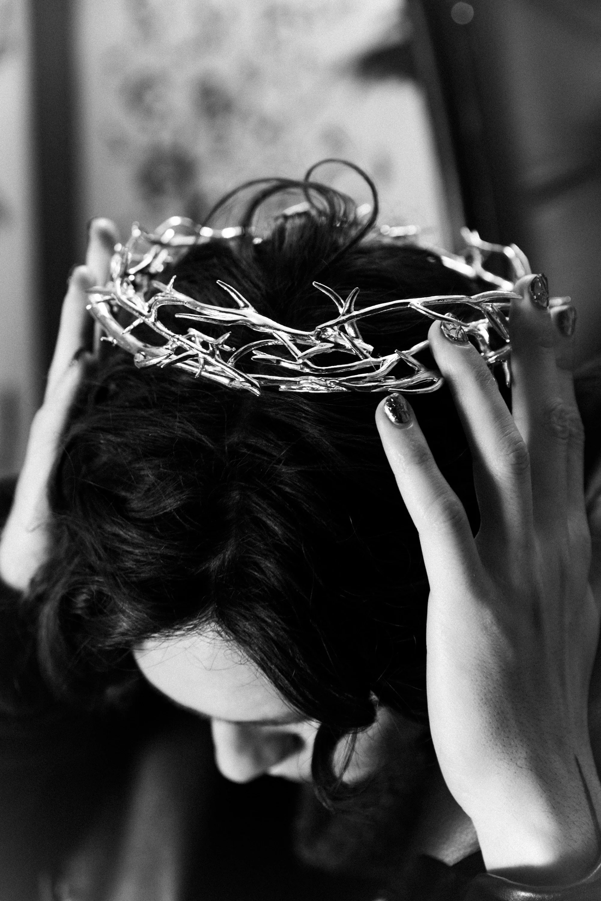 A person with dark hair is holding a decorative crown or headpiece with both hands on their head. The background is blurred and the photo is black and white.