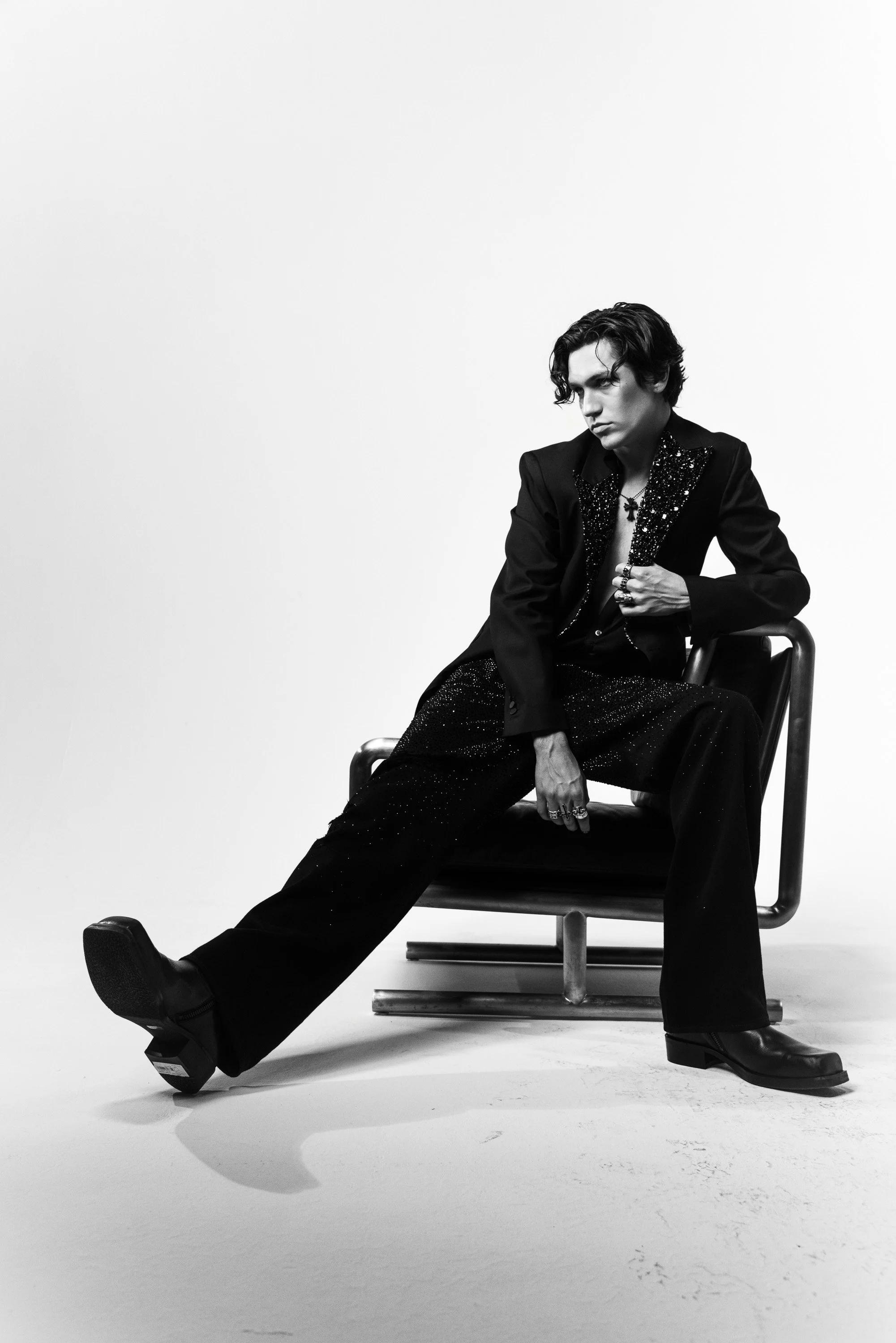 A black and white photo of a young man with wavy hair wearing a decorated blazer and loose-fitting pants, sitting on a chair in a minimalist studio with a plain white background.