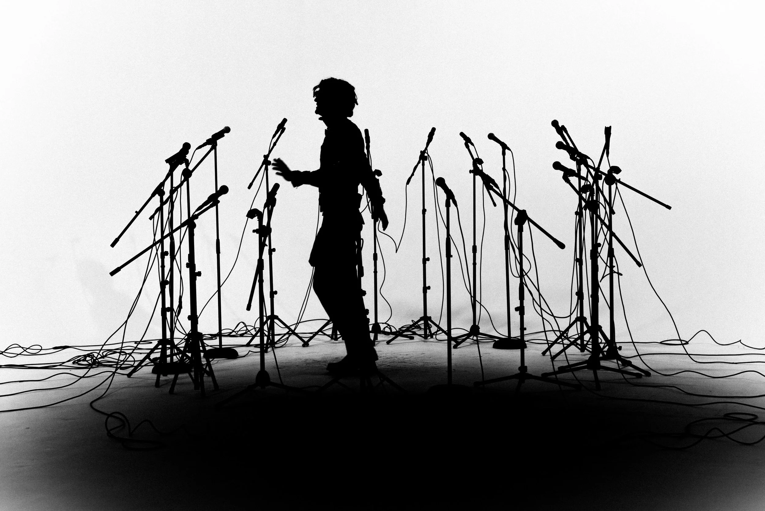 Silhouette of a person on stage surrounded by multiple microphone stands and cables, set against a plain background.