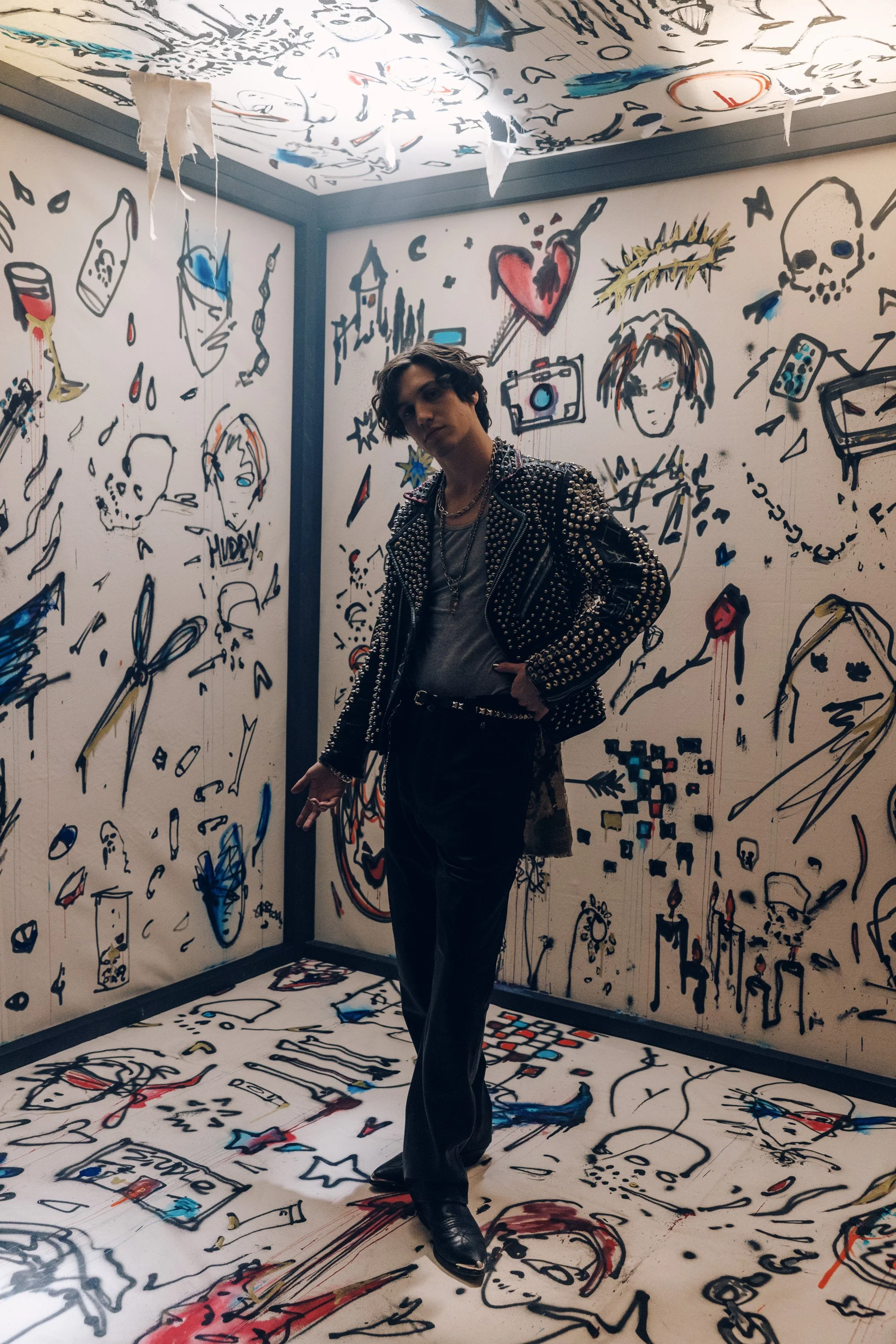 A young man with dark hair and tattoos wearing a black leather studded jacket, standing in an art installation room with walls and floor covered in black, red, blue, and yellow graffiti-style drawings and sketches, including faces, skulls, a heart, a