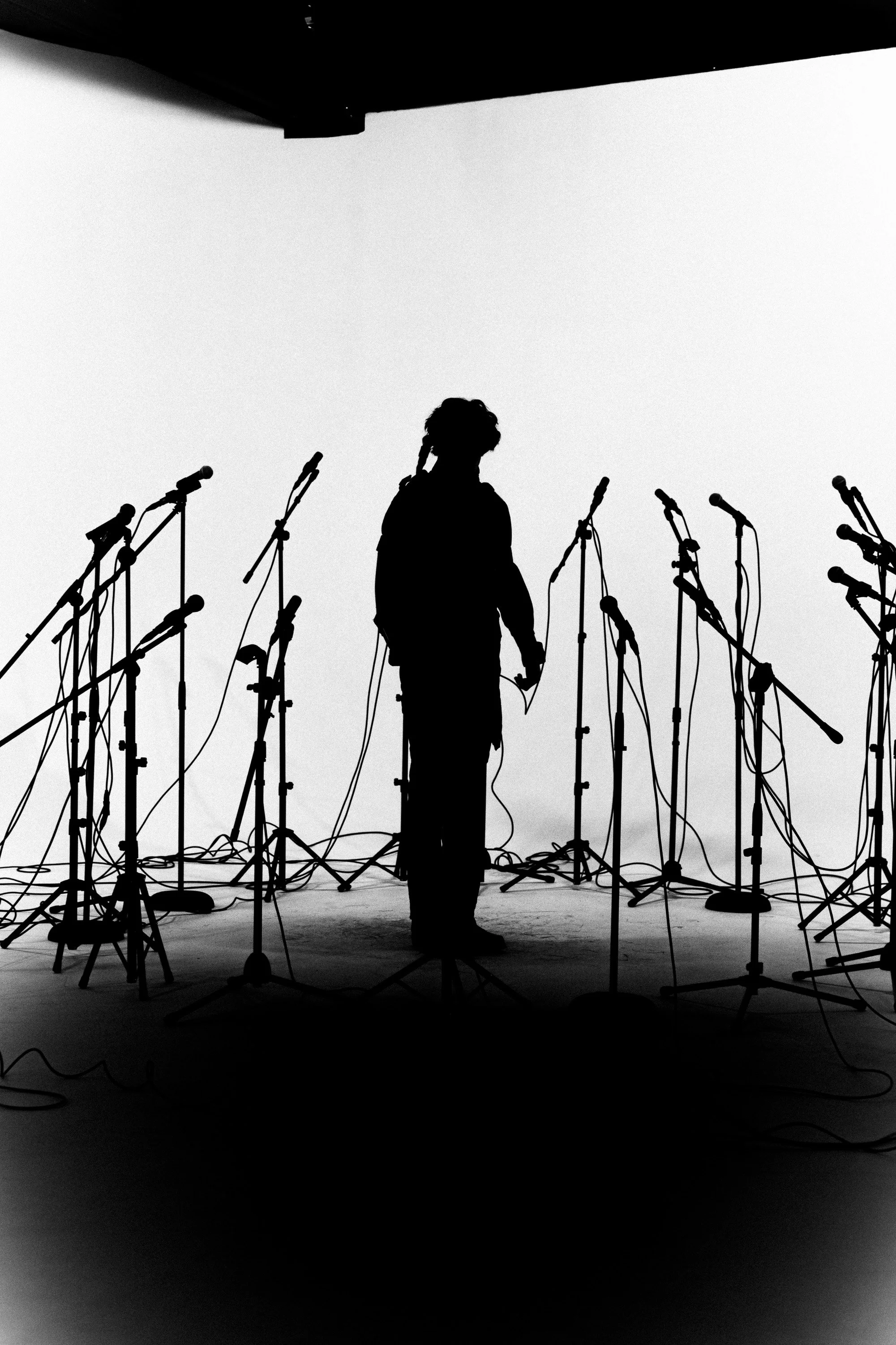 Black and white silhouette of a person standing among multiple microphones on stands, set against a plain light background.