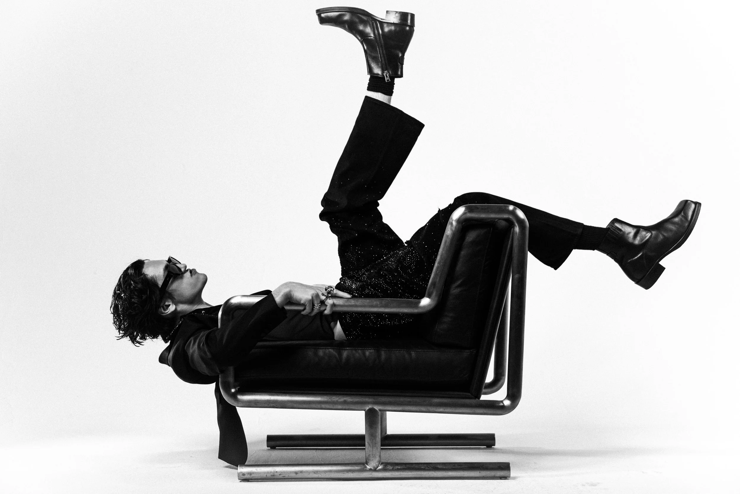 Black and white photo of a woman lying on a desk with her legs raised, wearing sunglasses, a shiny jacket, and black boots. The person appears to be in a playful pose.