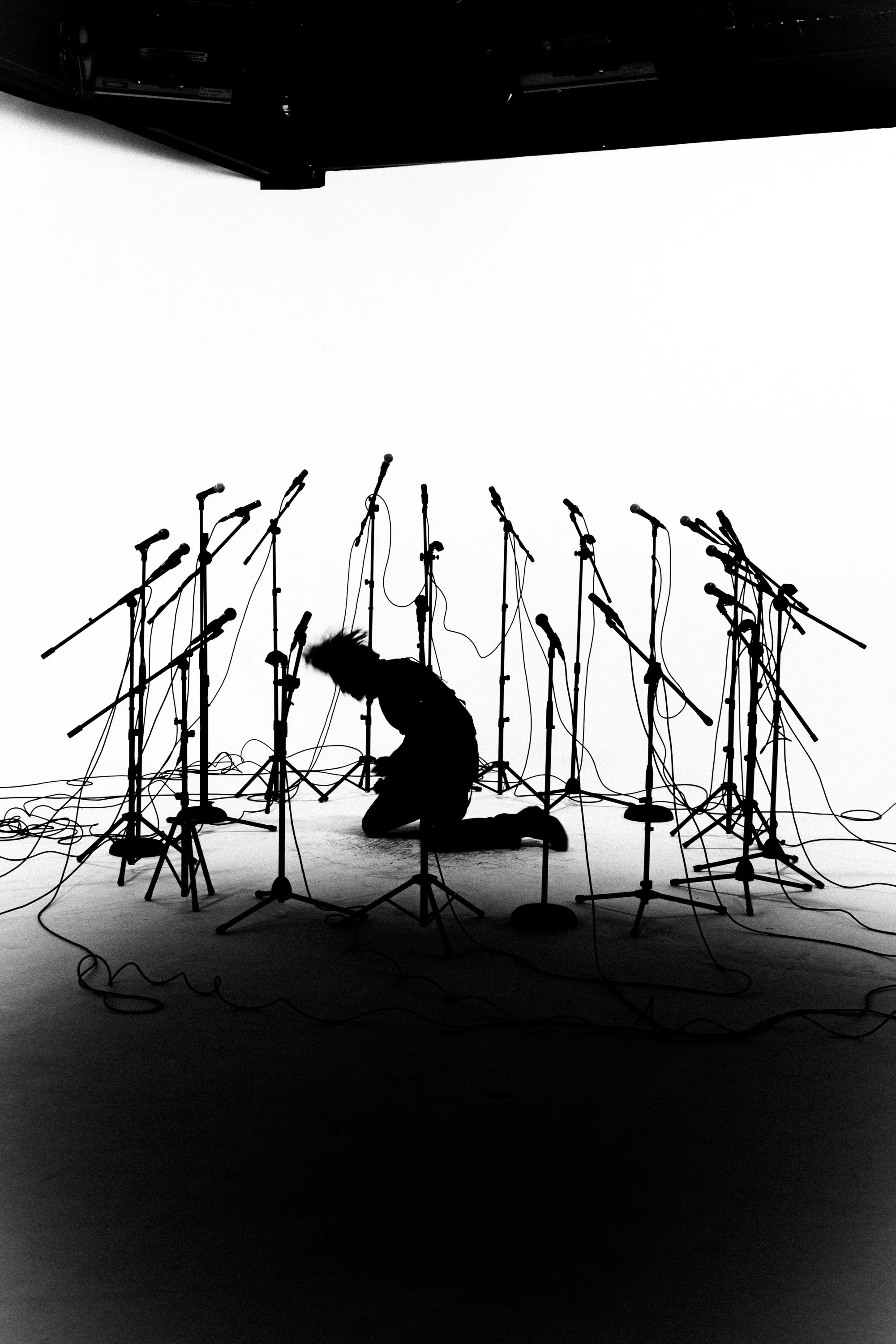 A person kneeling on the stage with multiple microphones set up on stands positioned in front of them, with cables on the floor, against a white background.