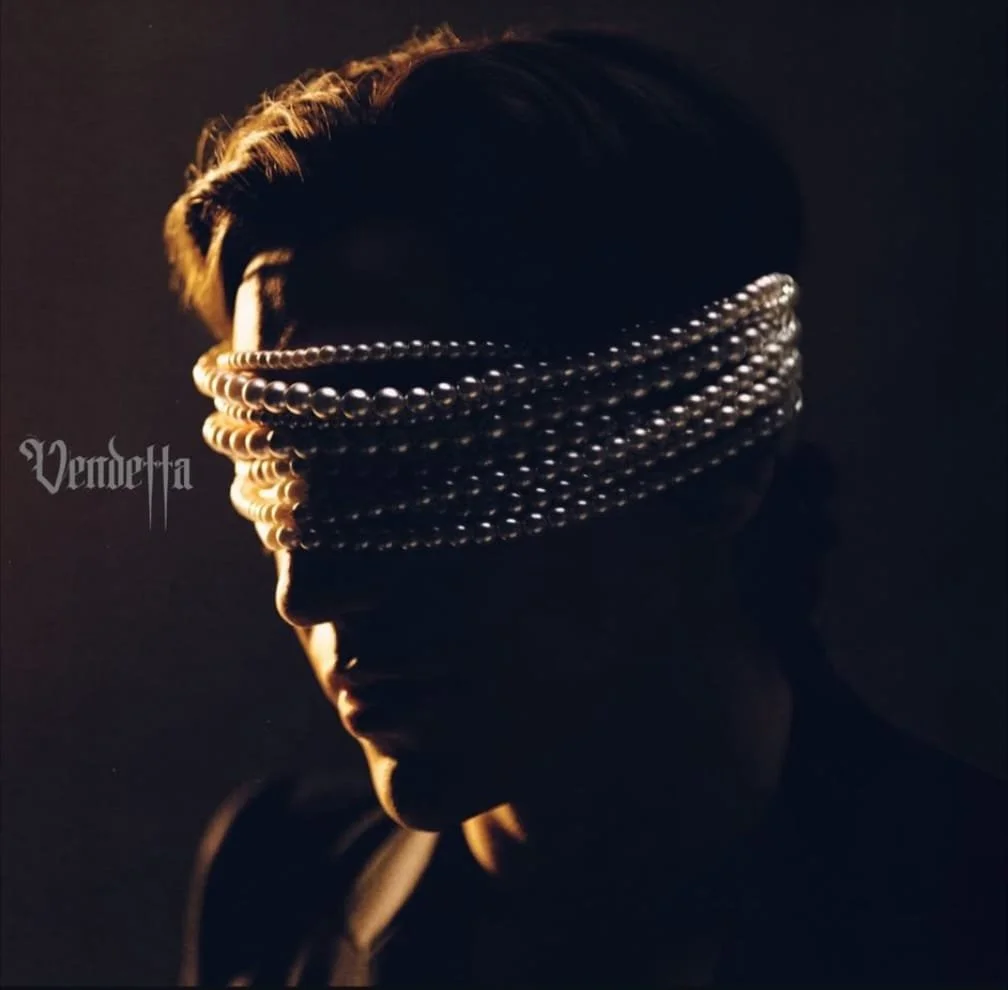 A person with dark hair styled upwards, partially illuminated by soft light, wearing multiple pearl necklaces around their forehead over eyes, against a dark background.