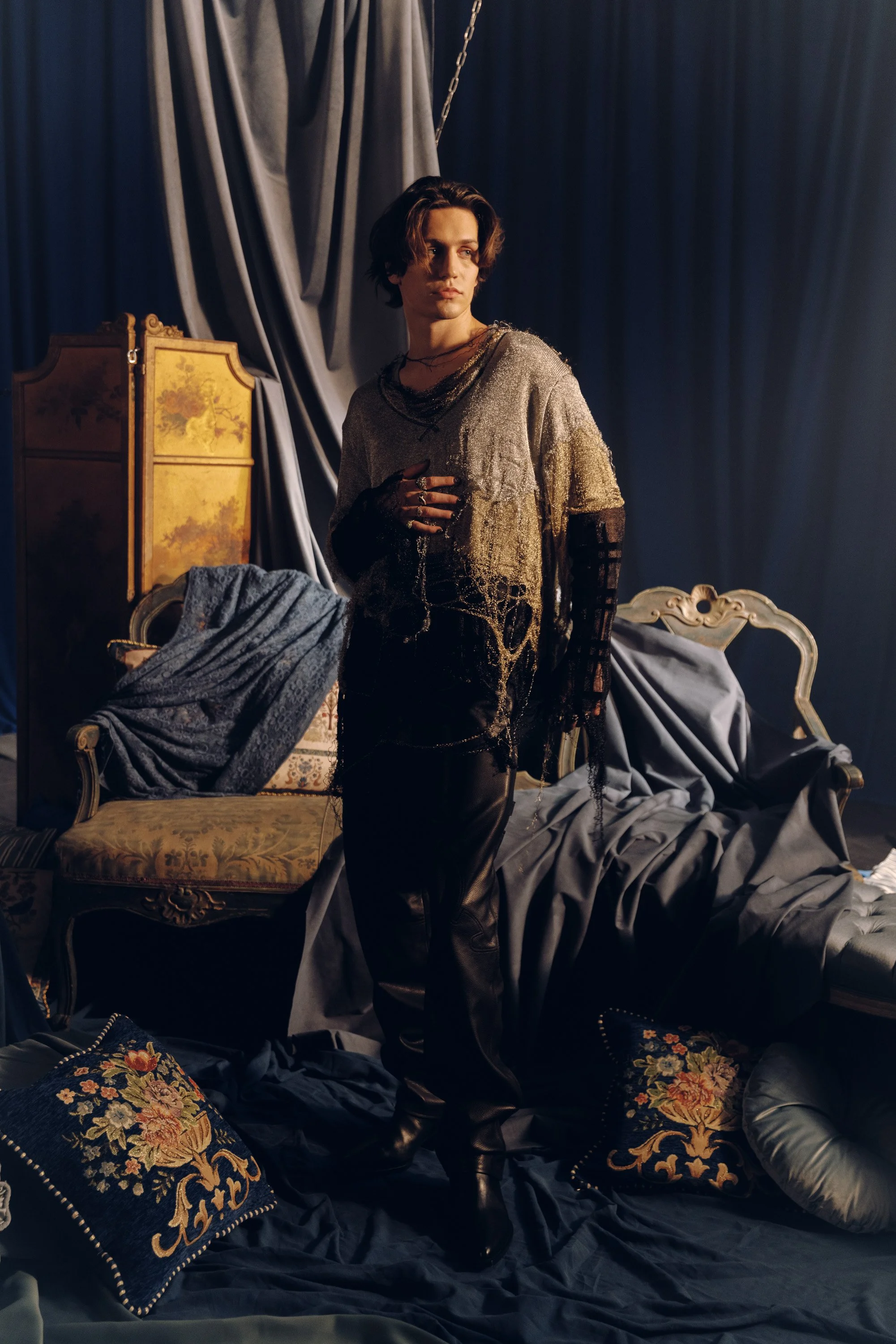 A person with short dark hair standing amidst a messy scene with various cushions, a chair, and a draped curtain in a dimly lit room, dressed in shiny and dark clothing.