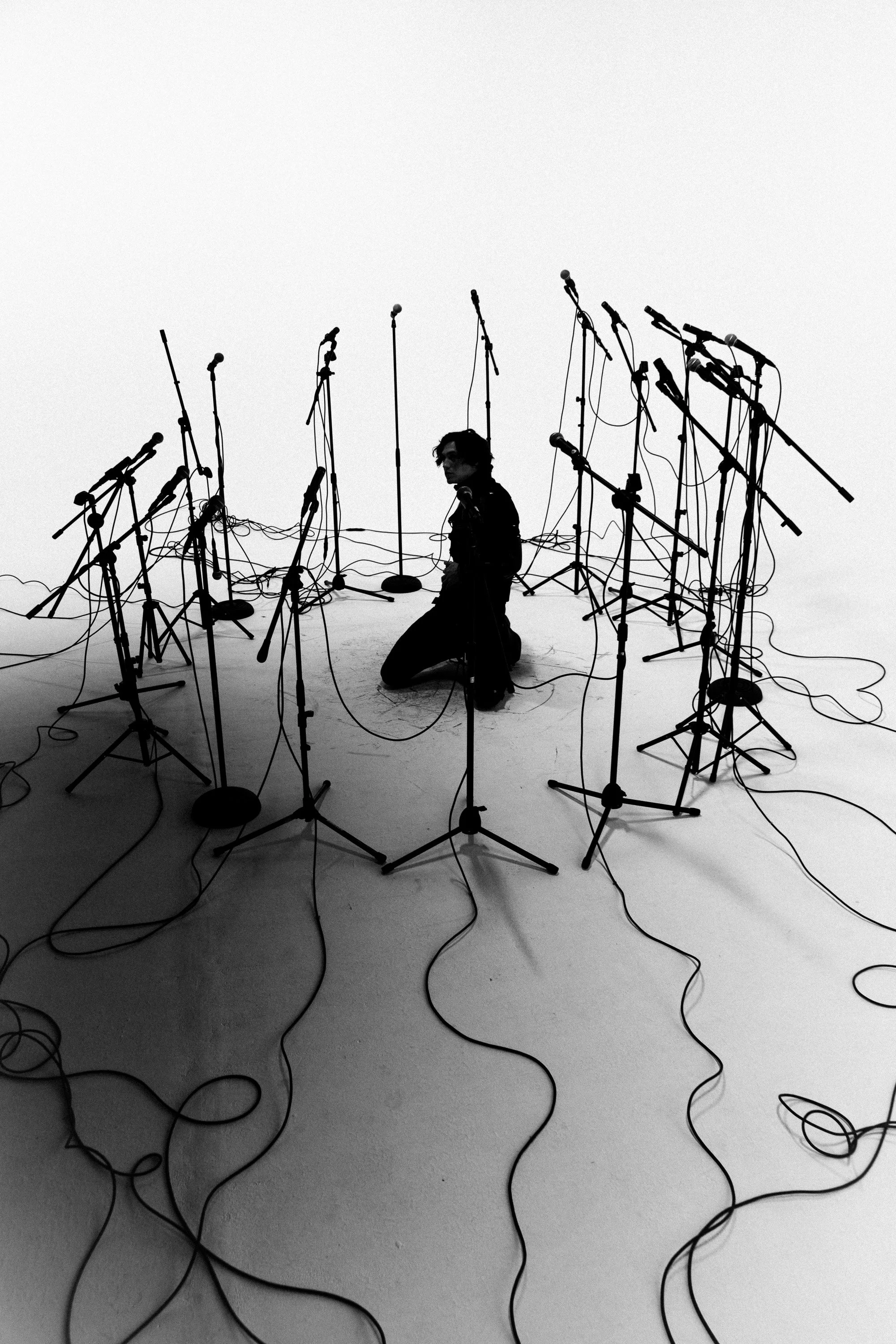 A black and white photo of a man kneeling on the floor, surrounded by numerous microphones on stands, with wires scattered across the floor and microphones positioned at various heights.