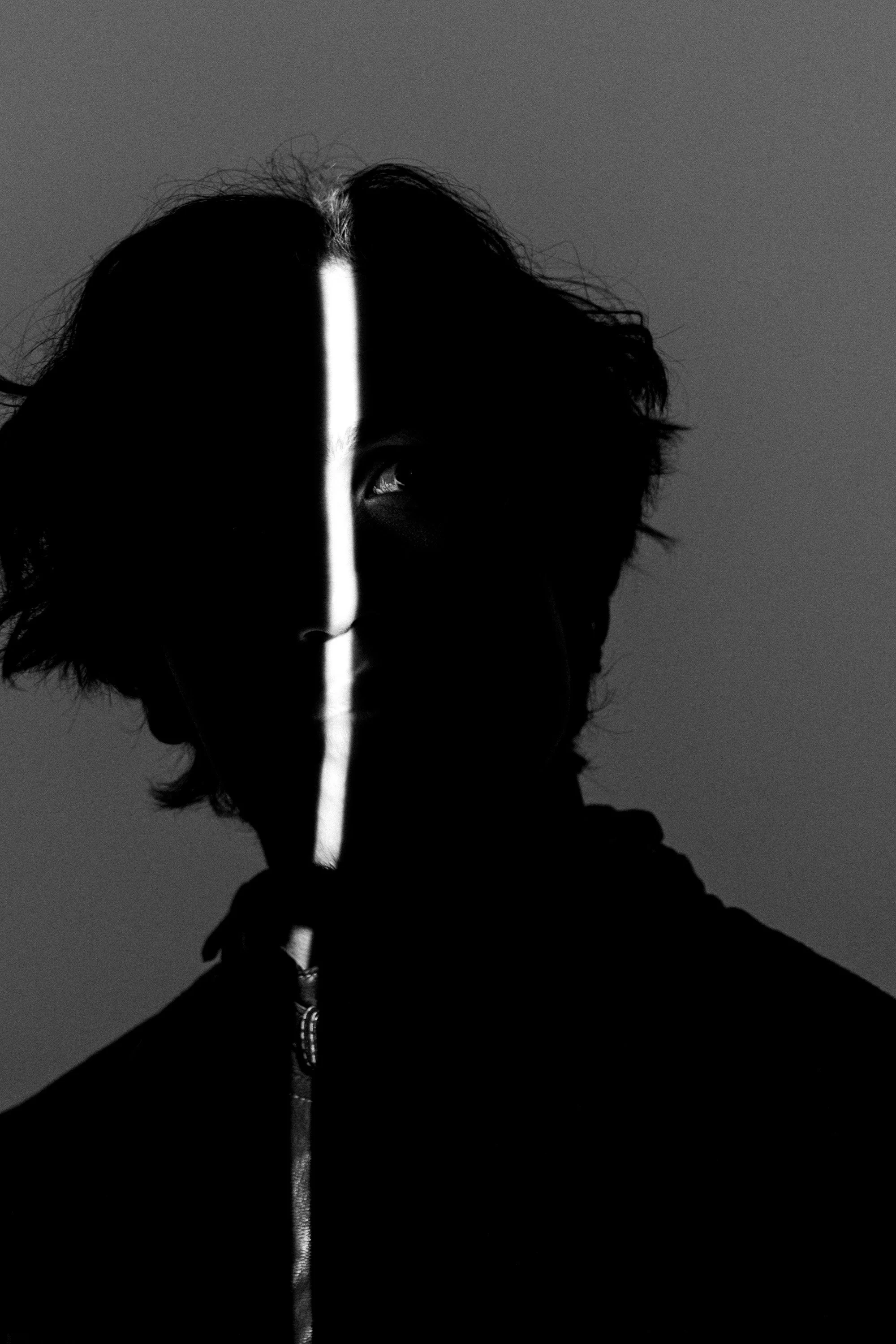 Black and white portrait of a person with dark, tousled hair, partially obscured face, and a vertical light streak across the face.