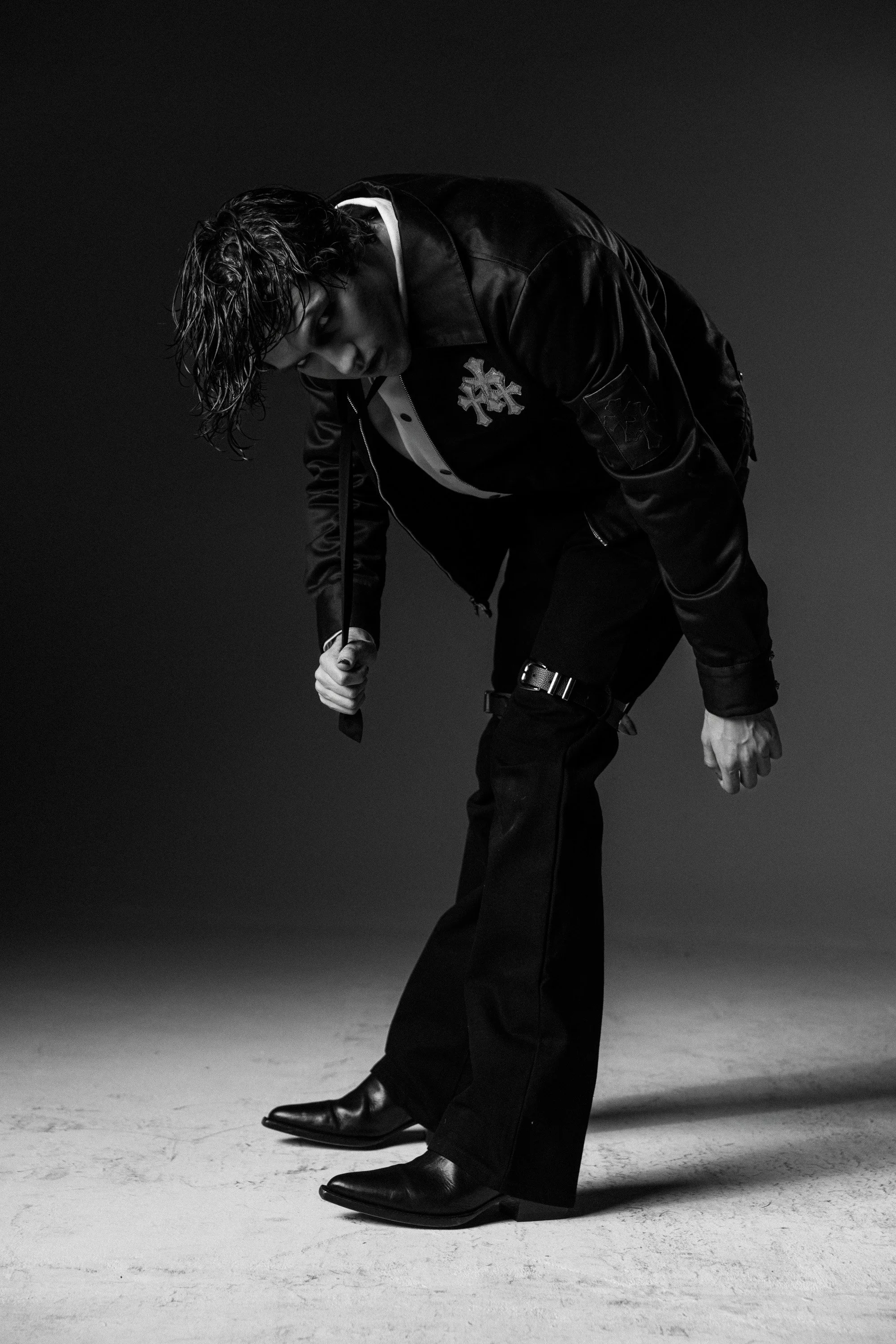 A man in black leather jacket and dress shoes, with messy hair, leaning forward with his eyes closed, against a dark background.