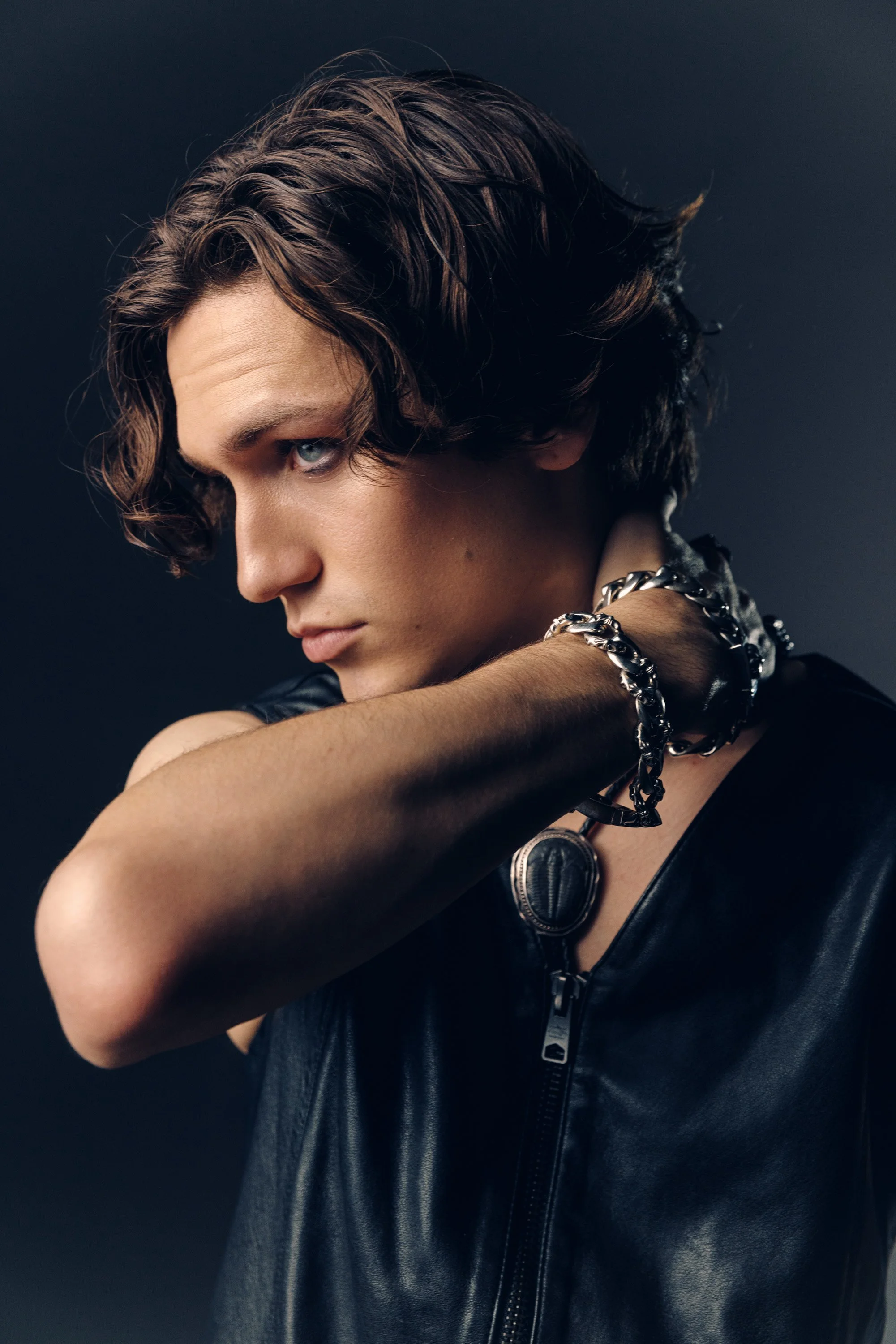 Close-up of a young man with wavy brown hair, wearing a black leather vest with a zipper, accessorized with silver chain bracelets and a watch, against a dark background.