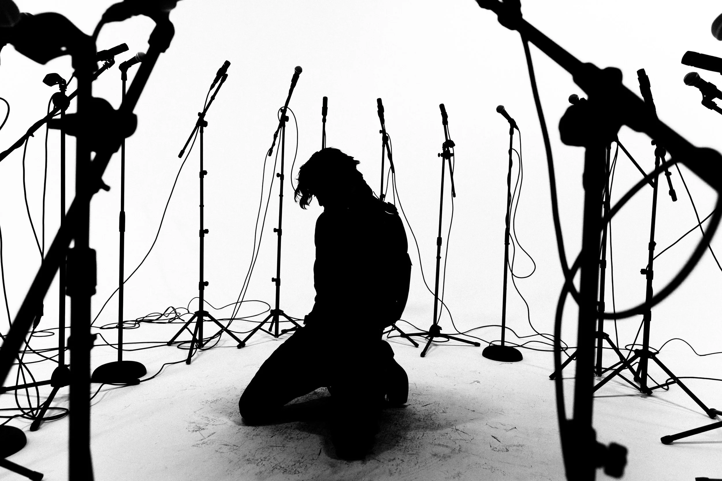 Silhouette of a person kneeling among multiple microphone stands on a stage or studio with a plain white background.
