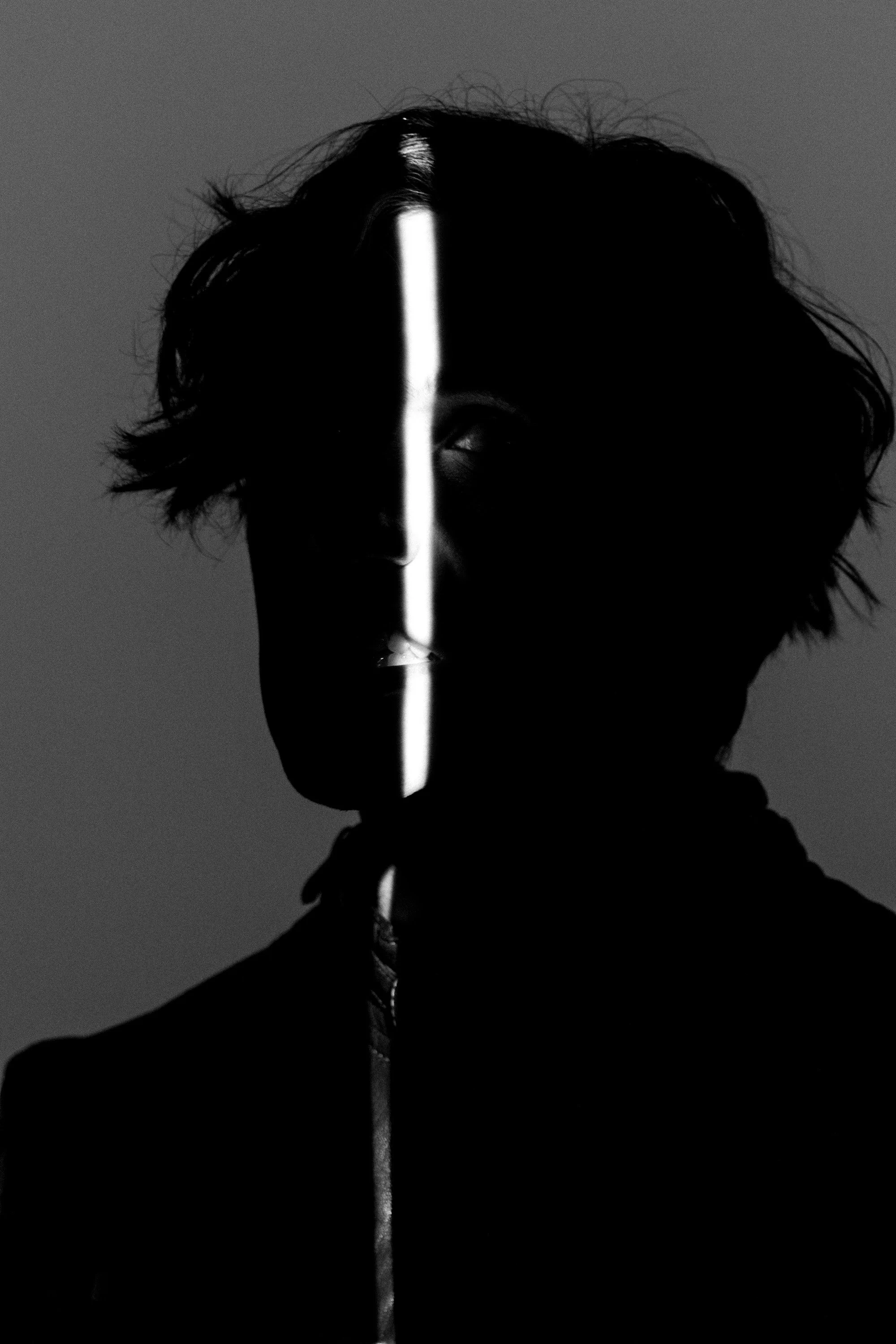 A black and white photo of a person's face mostly in shadow, with a vertical white light streak running vertically through the middle of the face, obscuring one eye and part of the nose.