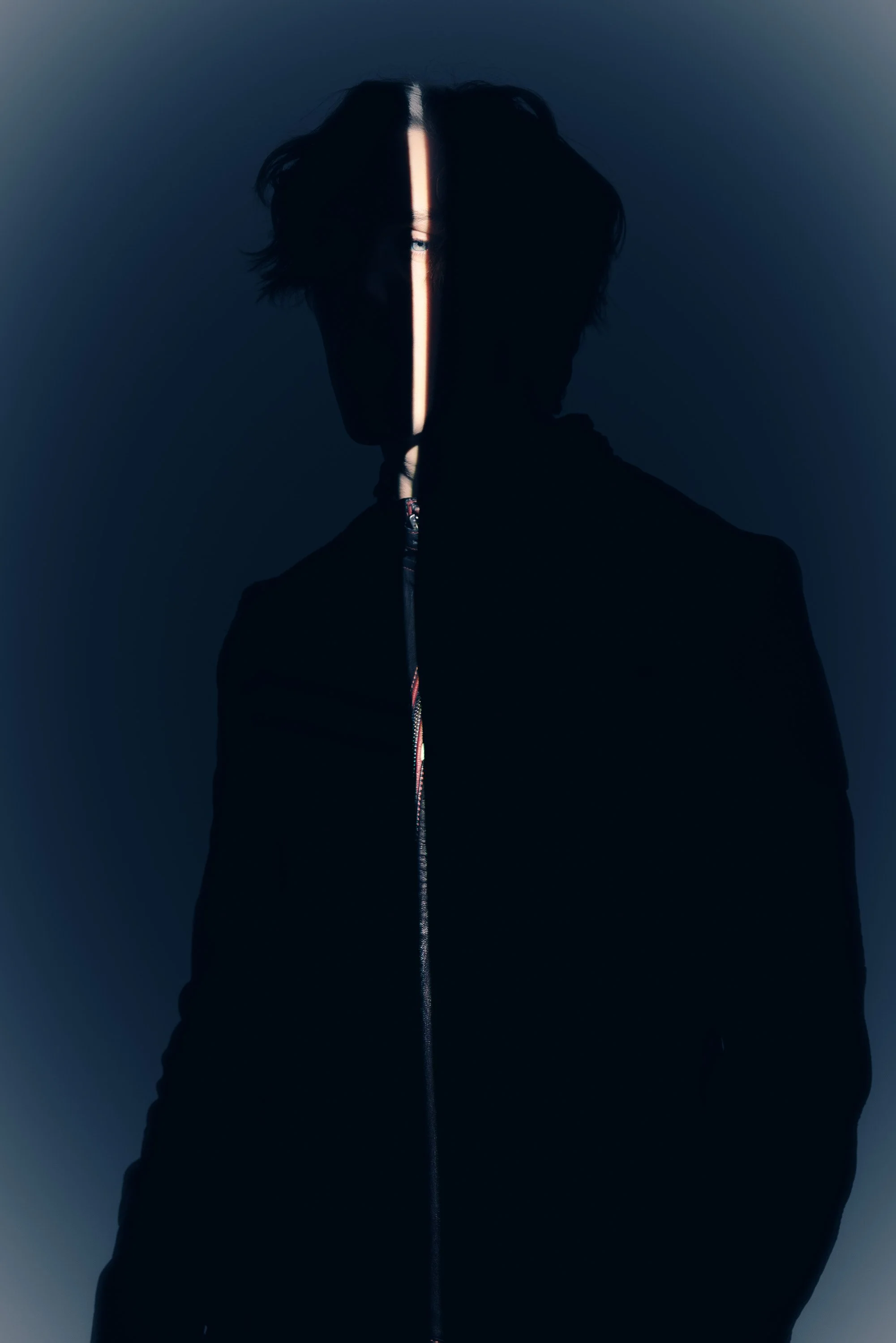 A person's silhouette with dark hair, facing forward, with a vertical strip of light crossing their face from forehead to chin, creating a mysterious and dramatic effect.