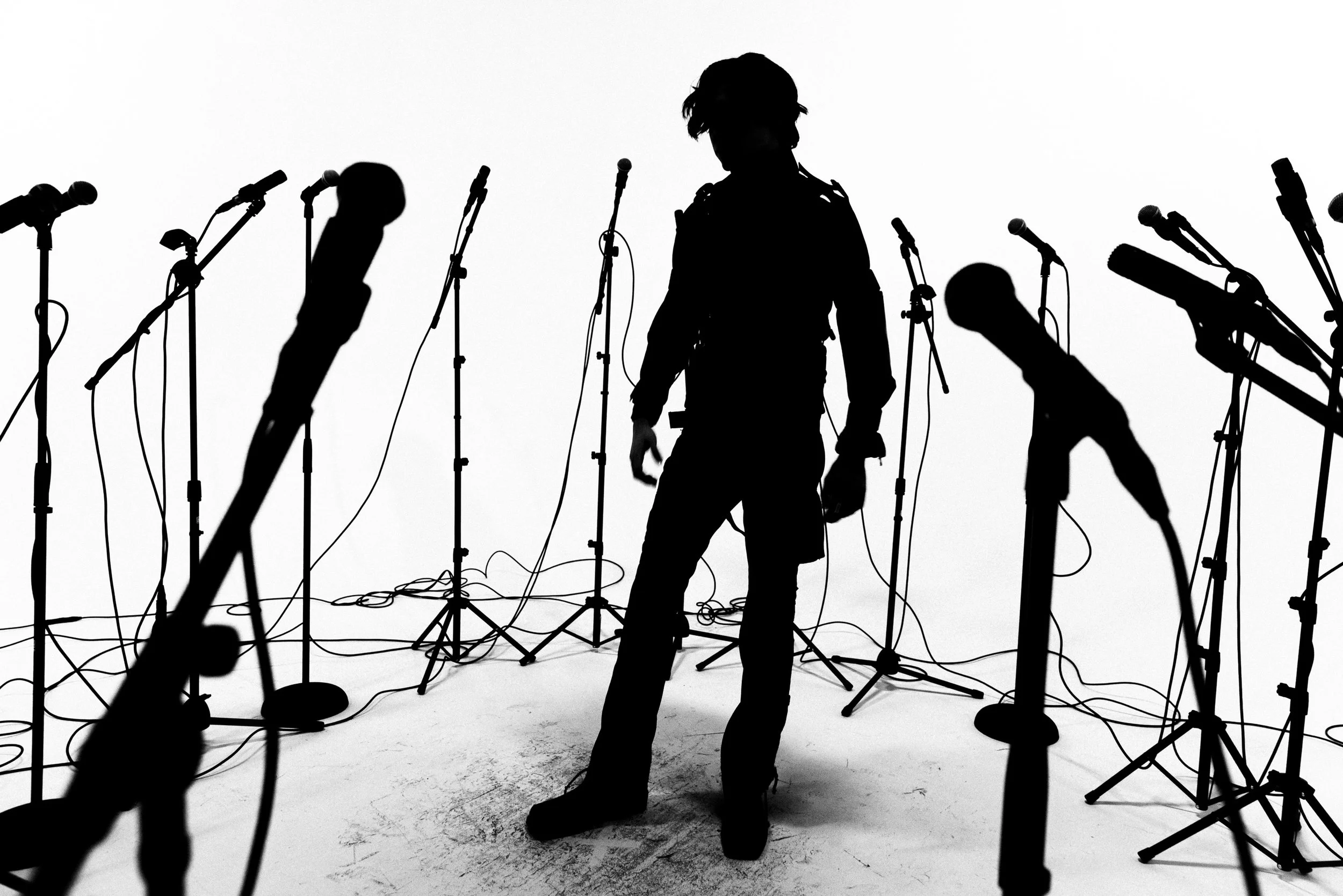 Silhouette of a person standing among multiple microphone stands on a plain background.
