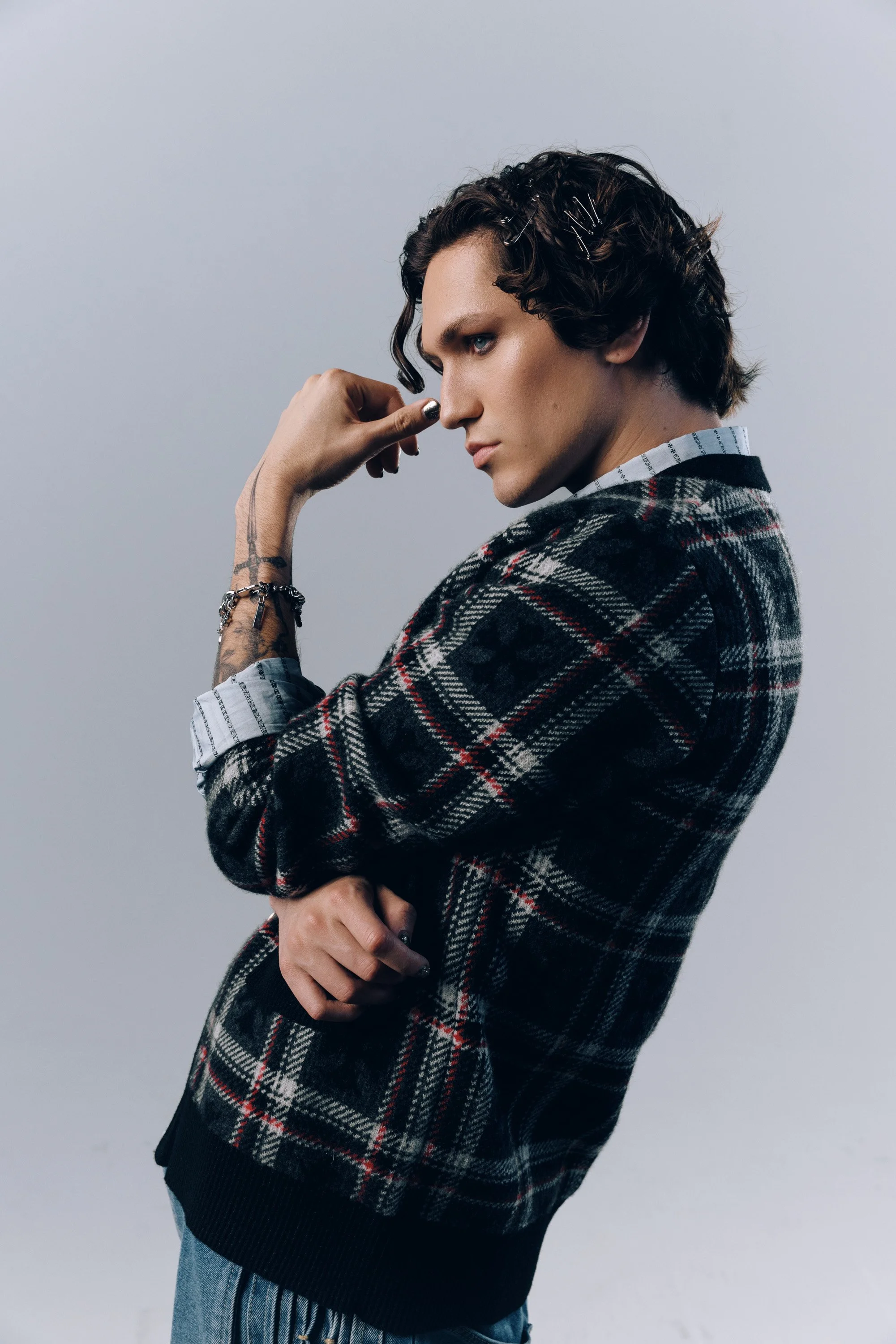 A young person with short, curly hair held back with hair clips, wearing a plaid sweater over a collared shirt, tattoos on the arm, and holding the nose with the hand, posed against a plain gray background.