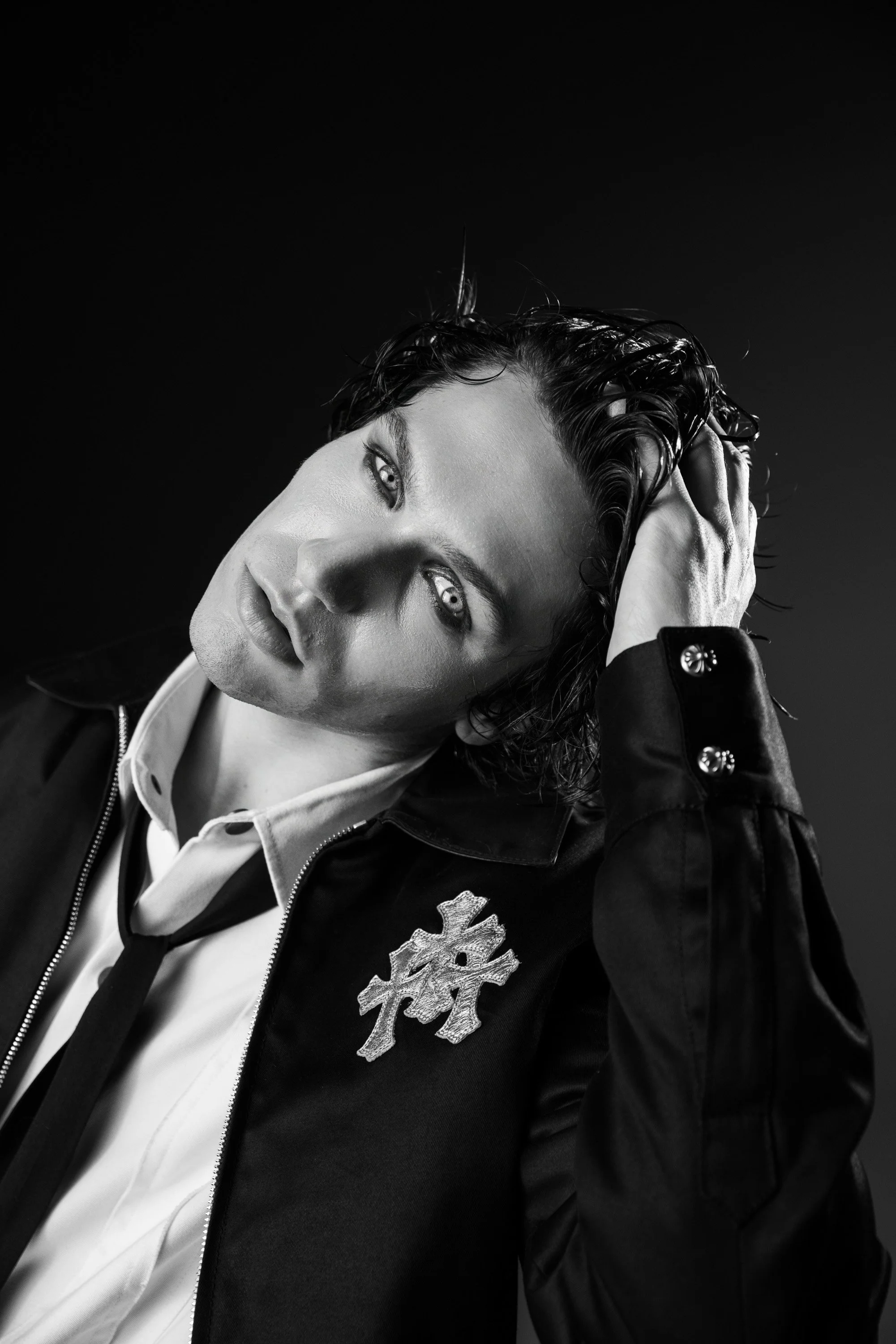 Black and white portrait of a woman with wet hair, wearing a jacket with snowflake patch, white shirt, and black tie, holding her head with one hand and looking into the distance.