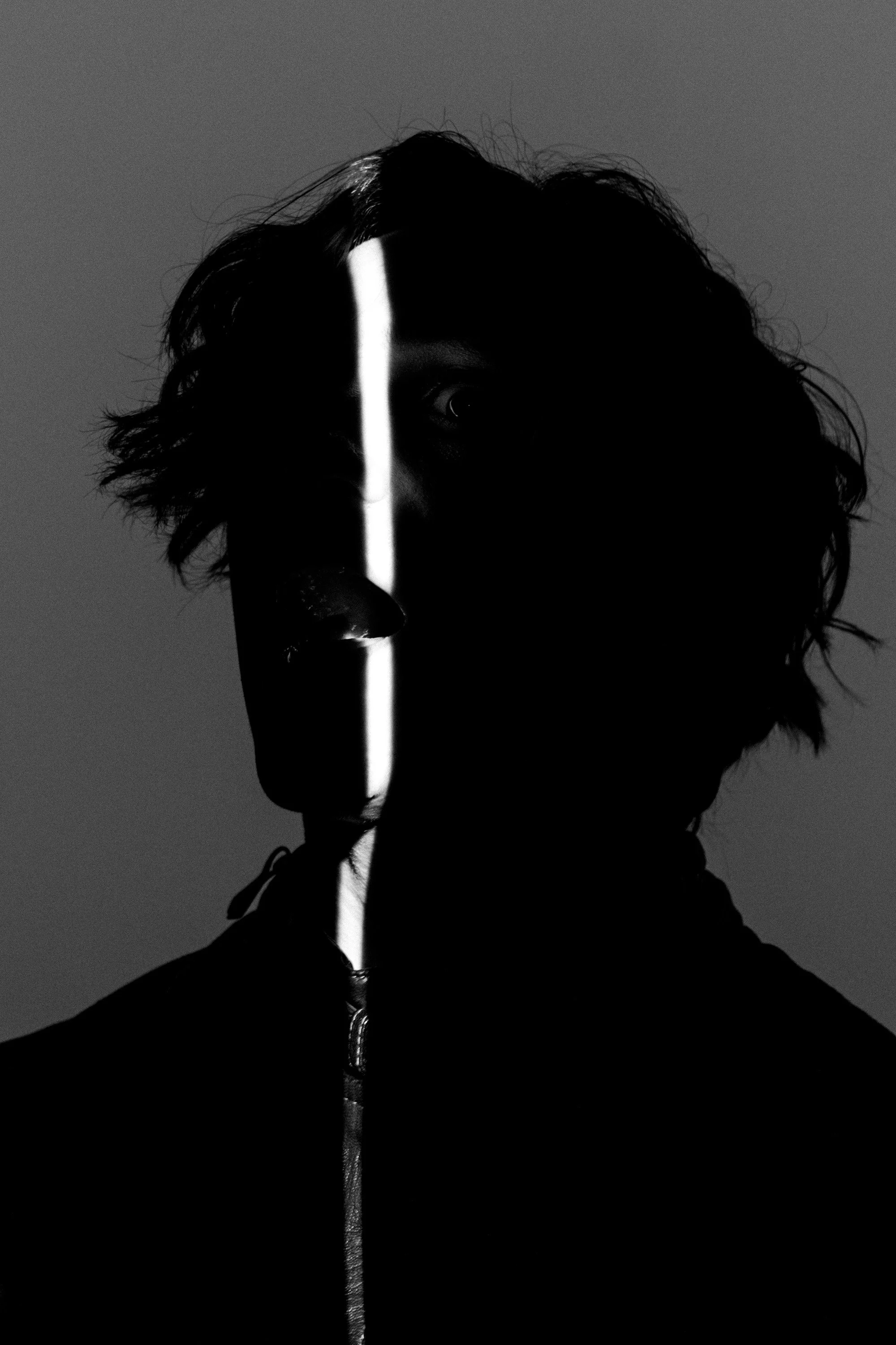 Black-and-white photo of a person with curly hair, with a narrow vertical strip of light illuminating their face from the nose down, creating a stark contrast against the dark background.