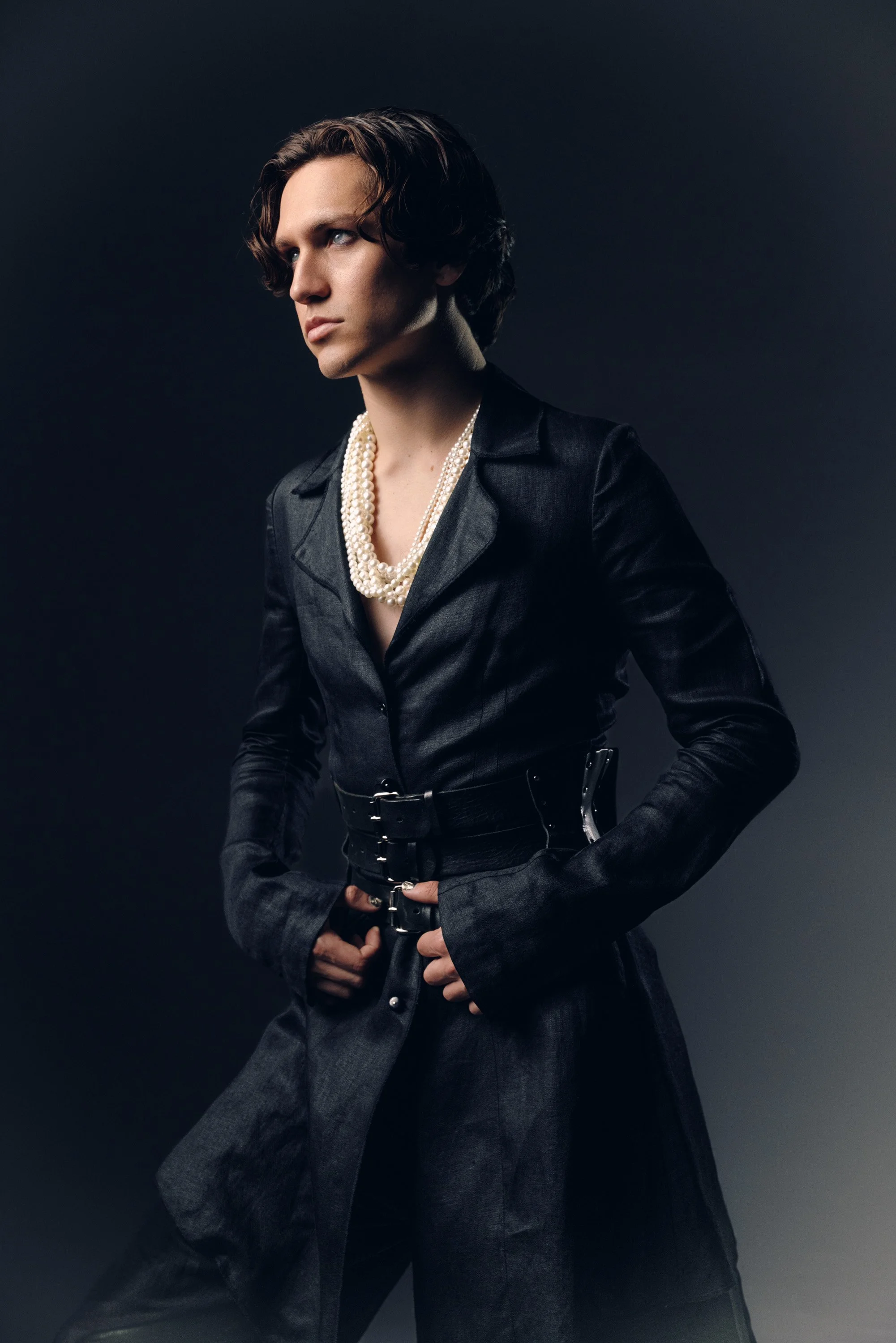 Portrait of a woman with short, wavy dark hair, wearing a black blazer with a pearl necklace, set against a dark background.