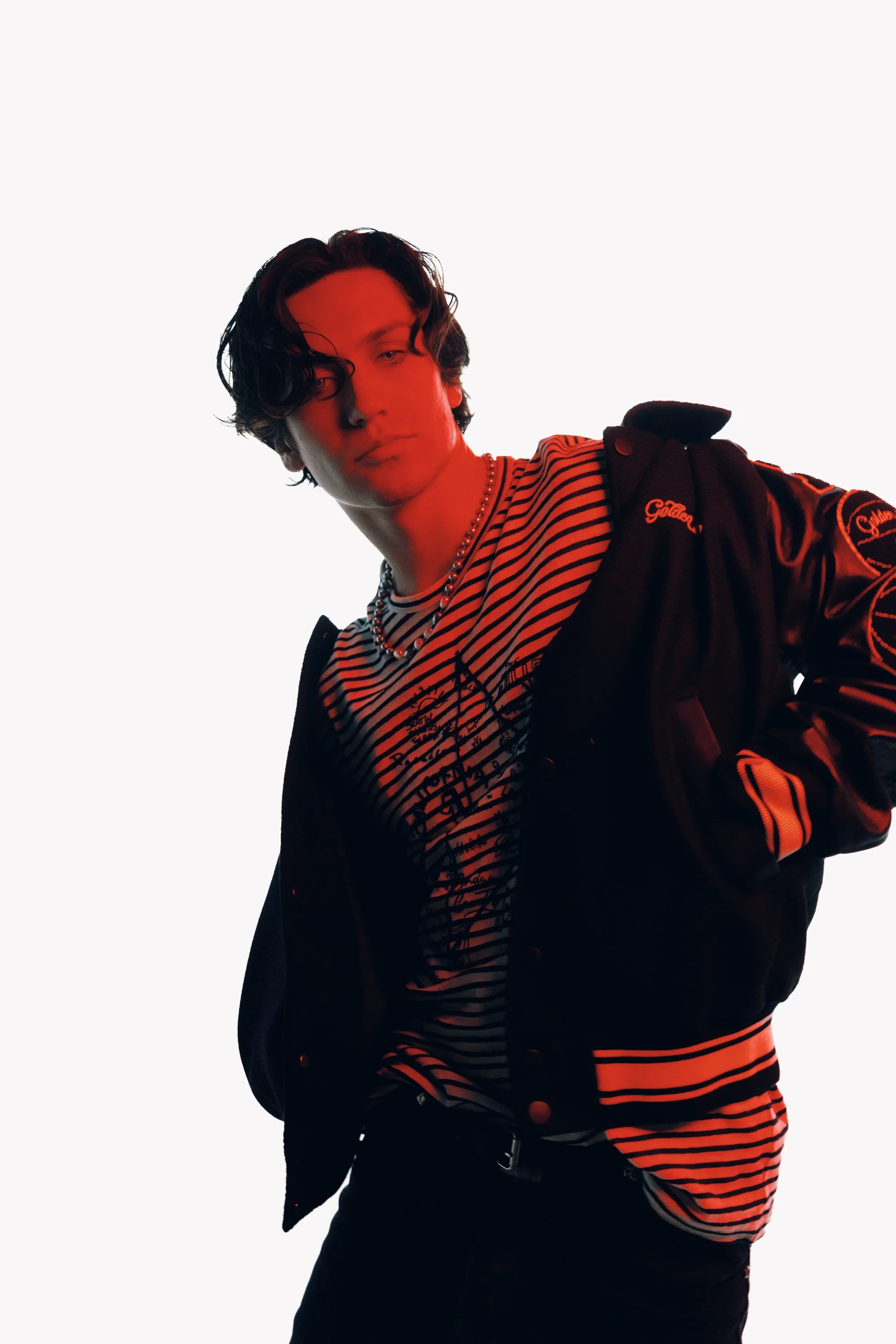 A young man with wavy dark hair wearing a striped shirt, a black jacket with red accents, and accessories, looking at the camera with a neutral expression against a plain white background.
