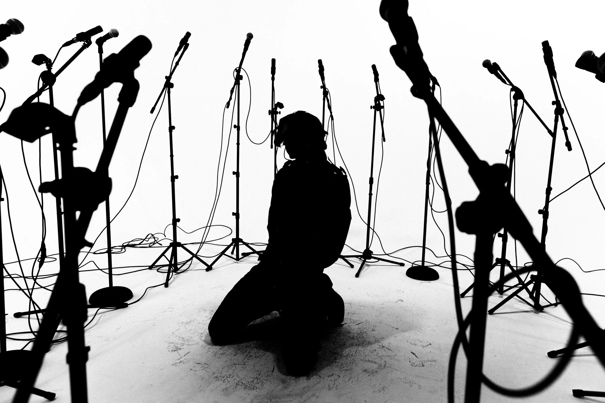 Silhouette of a person kneeling in front of multiple microphones on stands, with more microphones around them, in a studio or stage setting.