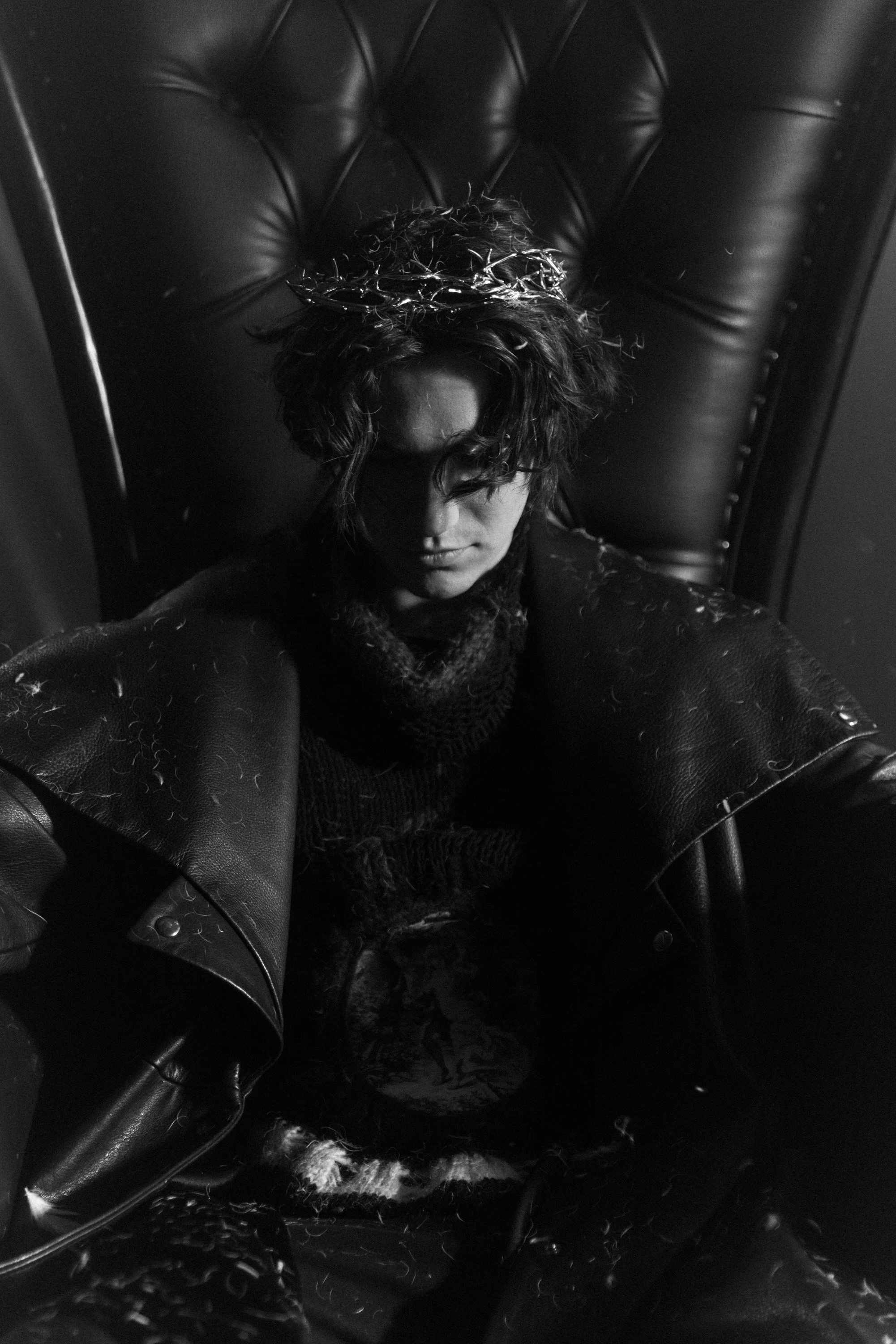 A person with curly hair sits on a leather chair or throne, wearing a crown made of barbed wire. The photo is in black and white, with the person's face partly obscured by their hair and the dark, textured surroundings.