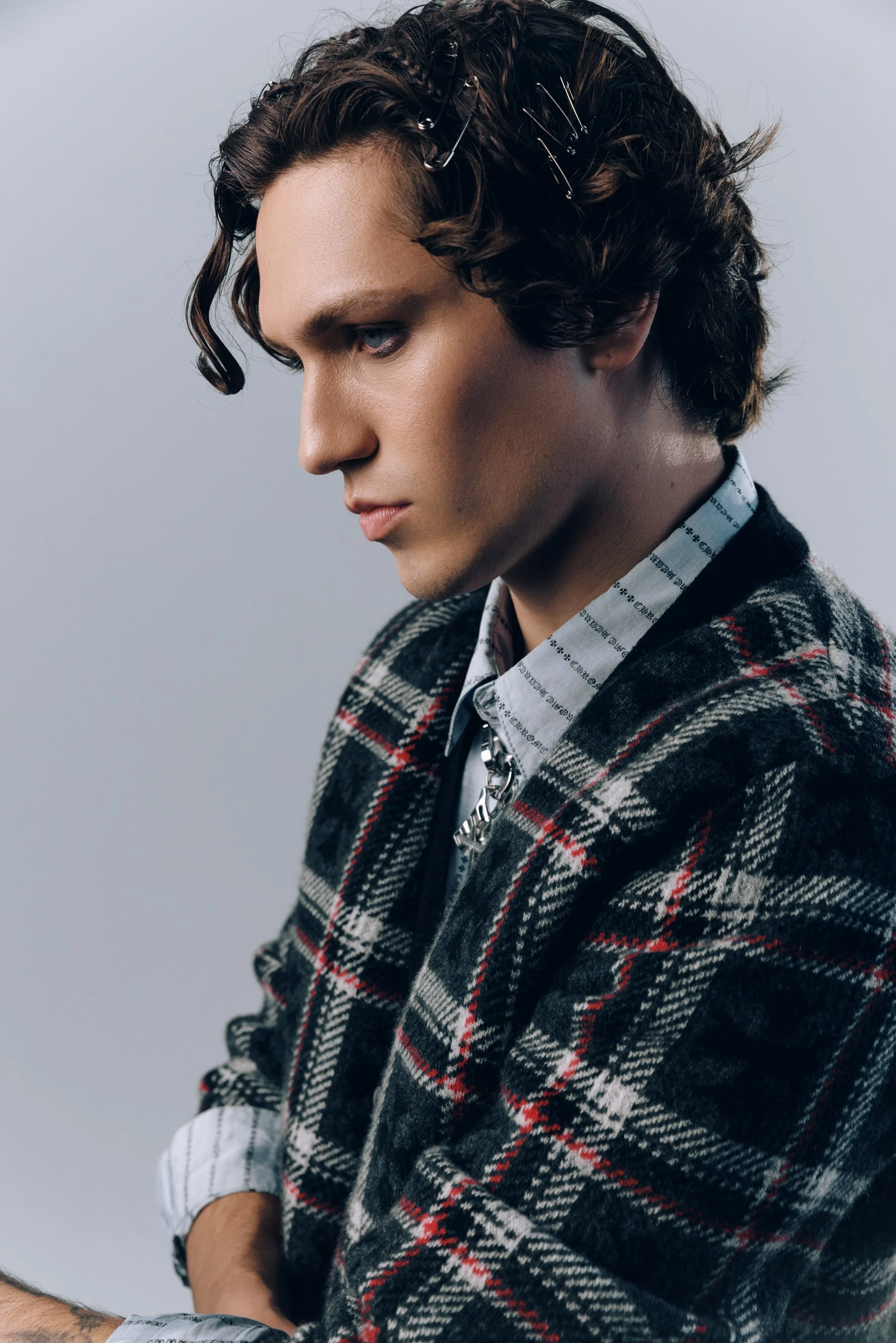A young man with dark, curly hair held back with hairpins, wearing a patterned blazer and a collared shirt with writing, looking downward with a serious expression against a plain gray background.