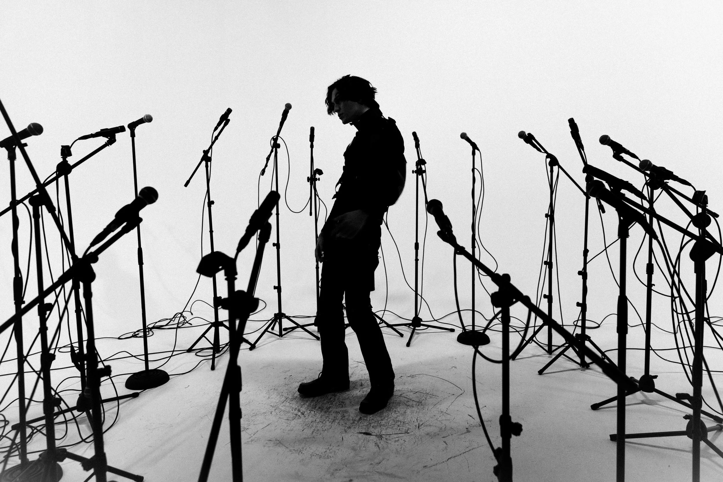 A person standing among numerous microphone stands in a studio setting with a plain white background.